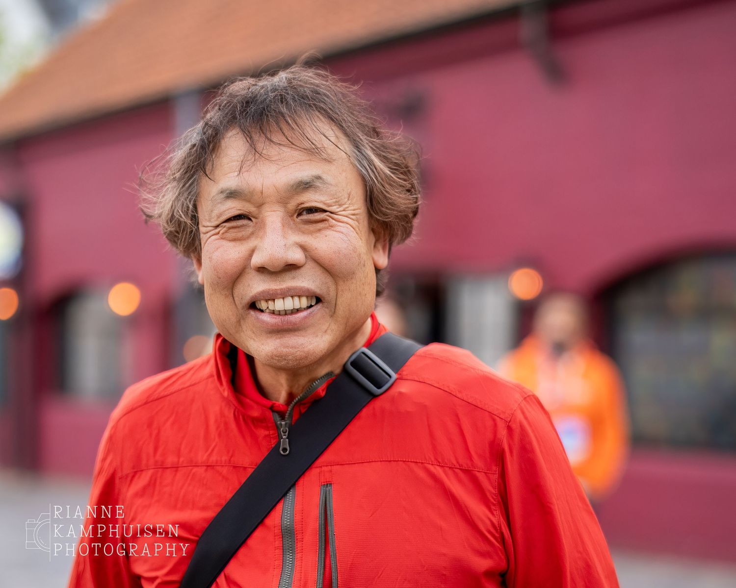 Man in rood jack lacht vriendelijk naar de camera, met een paarse gevel op de achtergrond.
