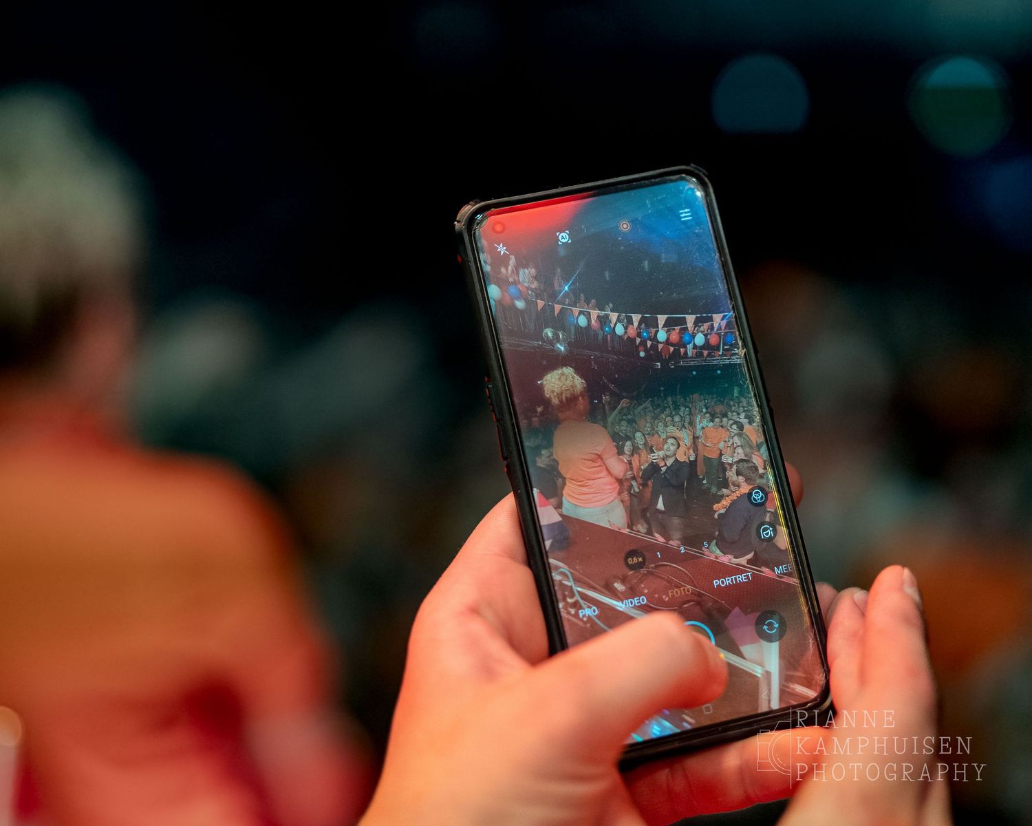 Close-up van een smartphone waarop een optreden wordt vastgelegd, met een dansend publiek en feestverlichting in beeld.