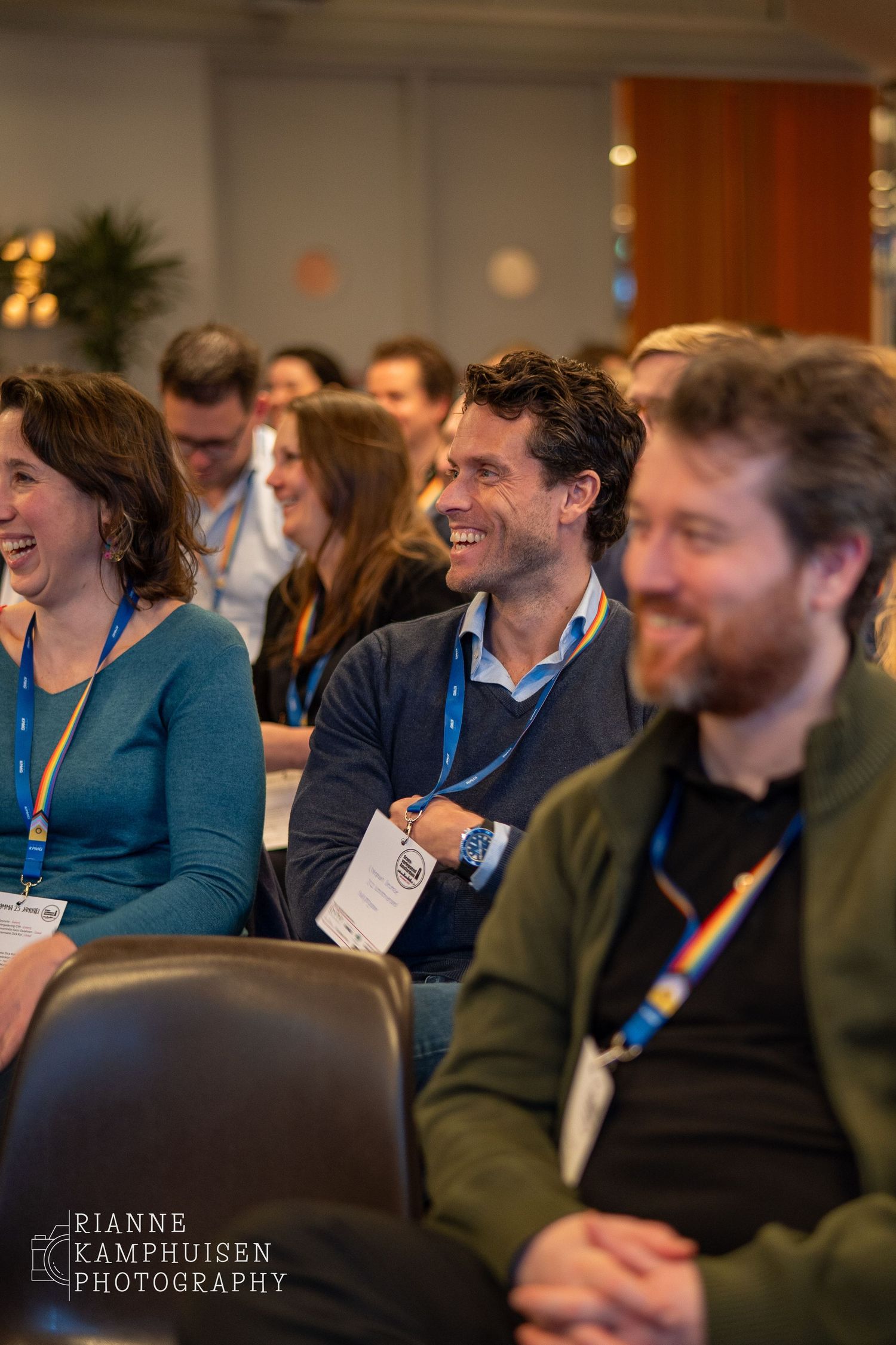 Publiek tijdens een evenement lacht en luistert aandachtig, met focus op een rij enthousiaste deelnemers met naambadges.