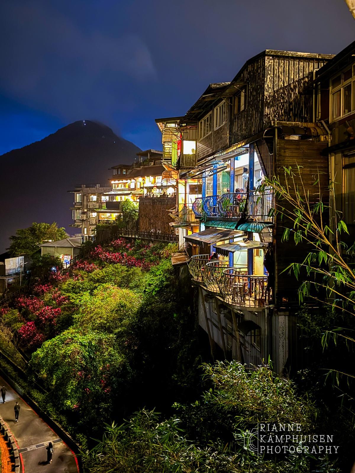 Traditionele houten huizen verlicht in de avond, tegen een berglandschap met kleurrijke bloemen op de voorgrond. Jiufen, Taiwan