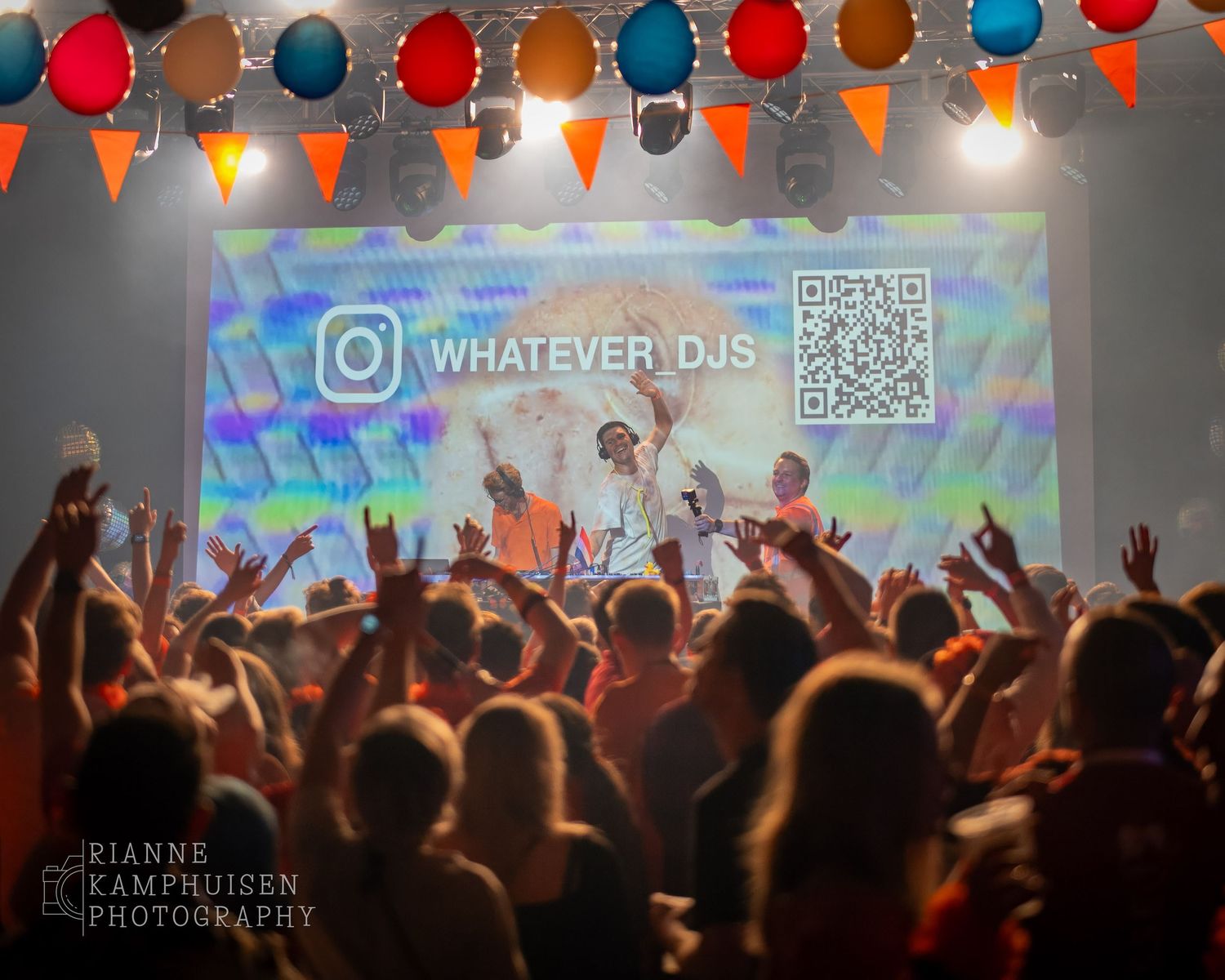 DJ's treden op voor een uitzinnig publiek onder kleurrijke feestverlichting, met projectie van “WHATEVER DJS” en een QR-code op het scherm.