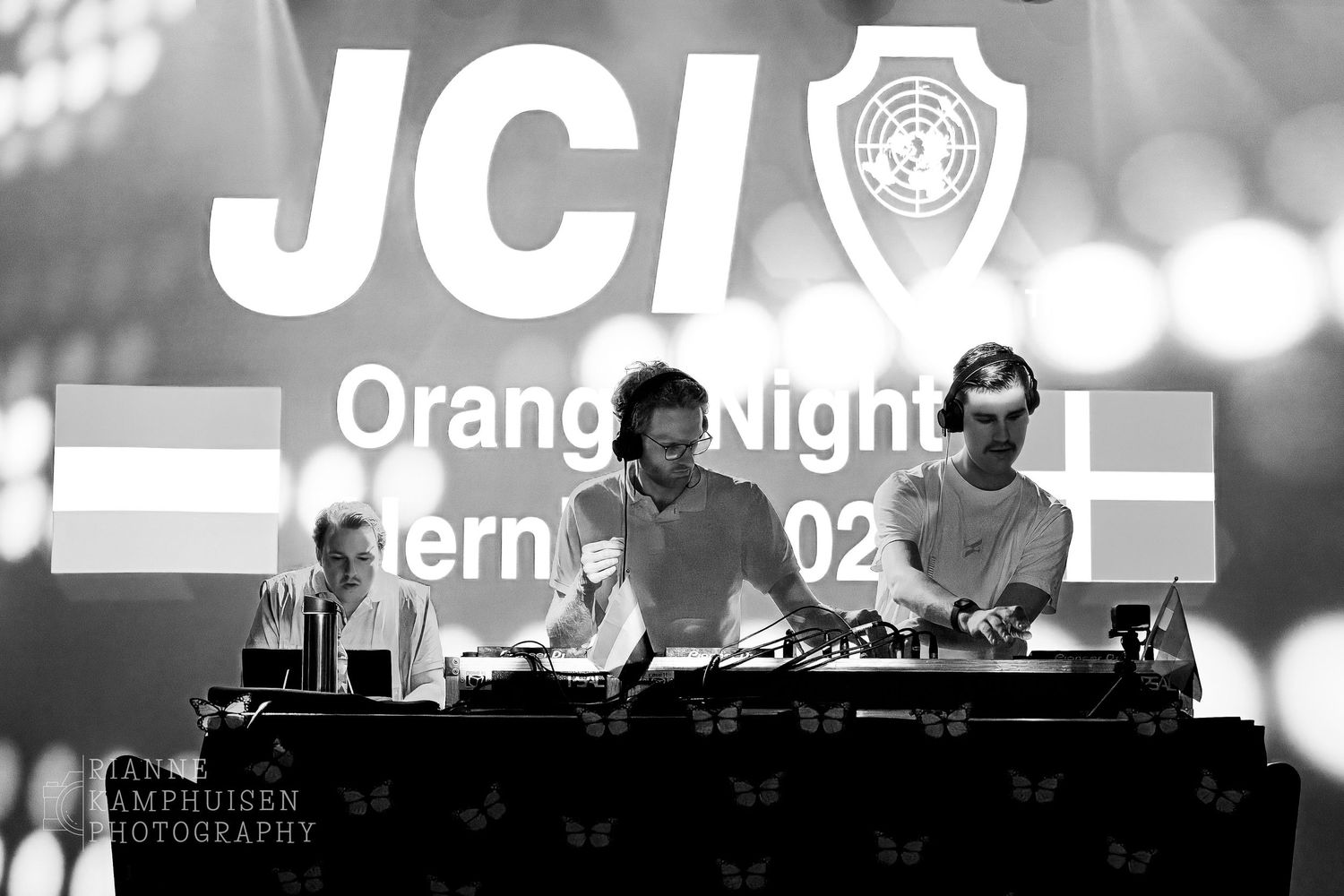 Drie DJ's draaien tijdens JCI Orange Night, met groot logo op achtergrond en sfeervolle lichtshow. zwart wit foto
