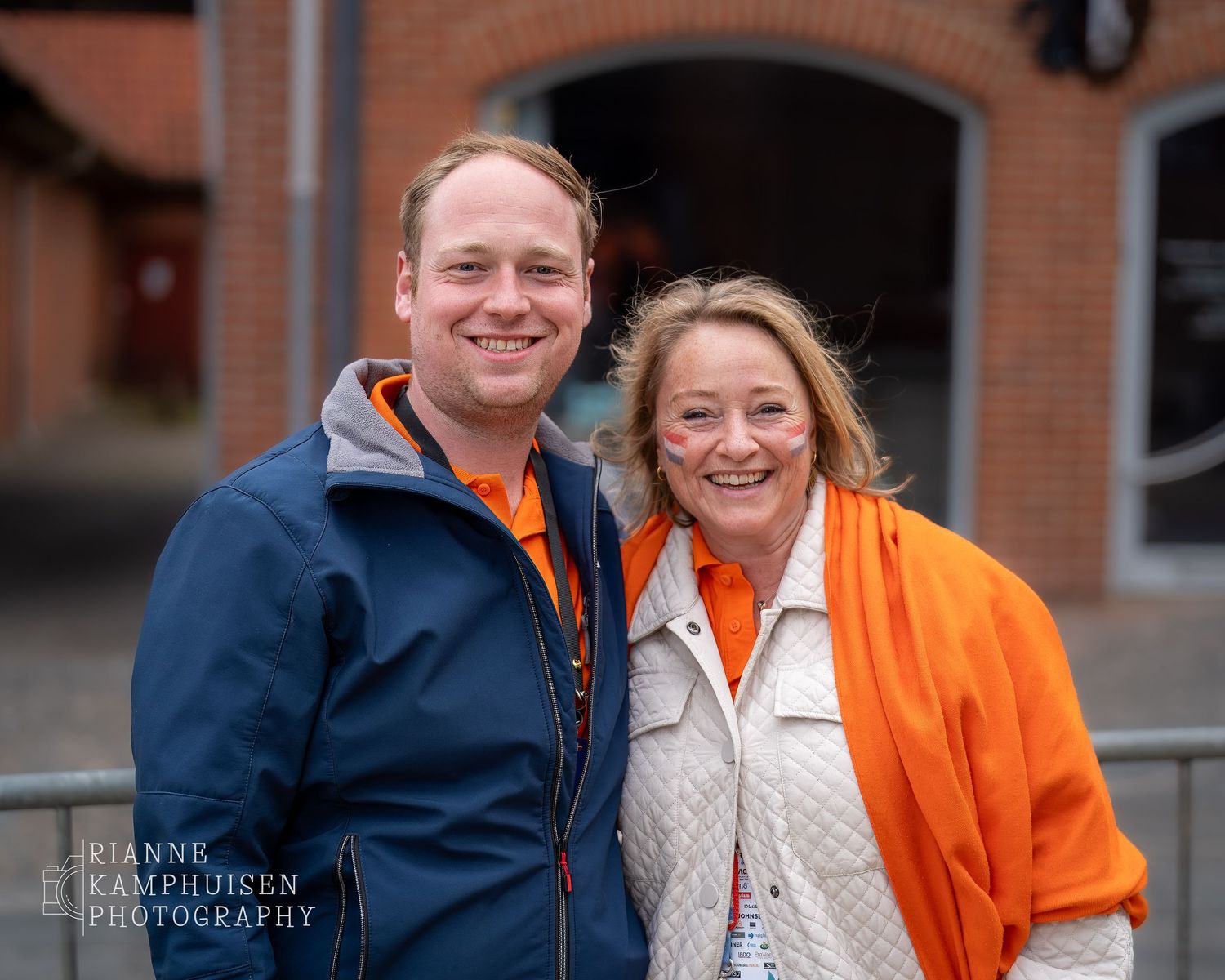 Twee mensen in oranje en blauw poseren lachend samen buiten tijdens een evenement.