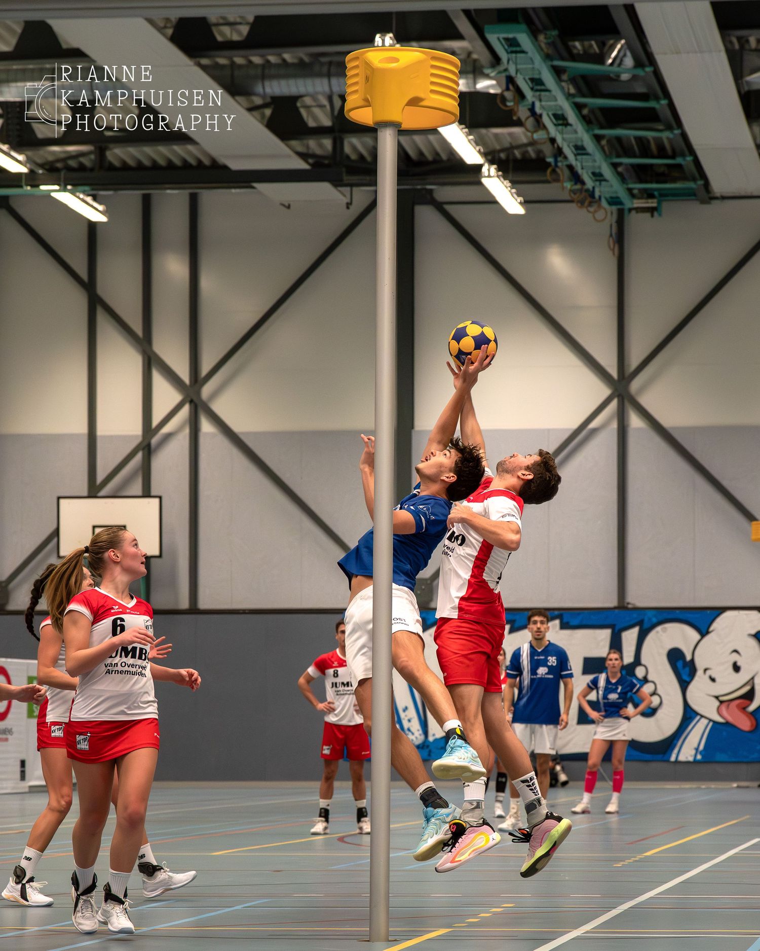 Twee korfbal spelers springen bij de korf omhoog, de aanvaller probeert te scoren, de verdediger wil met  zijn hand de bal blokken.