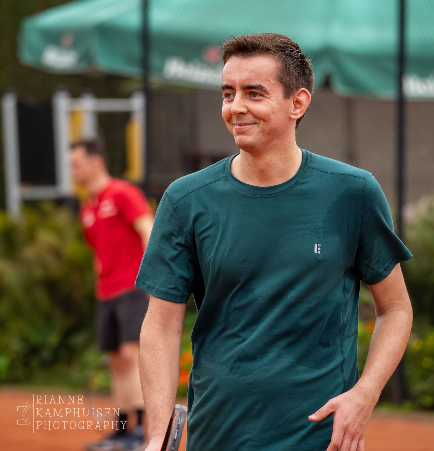 Tennisser in donkergroen shirt loopt tevreden over de baan, met lichte glimlach op zijn gezicht.
