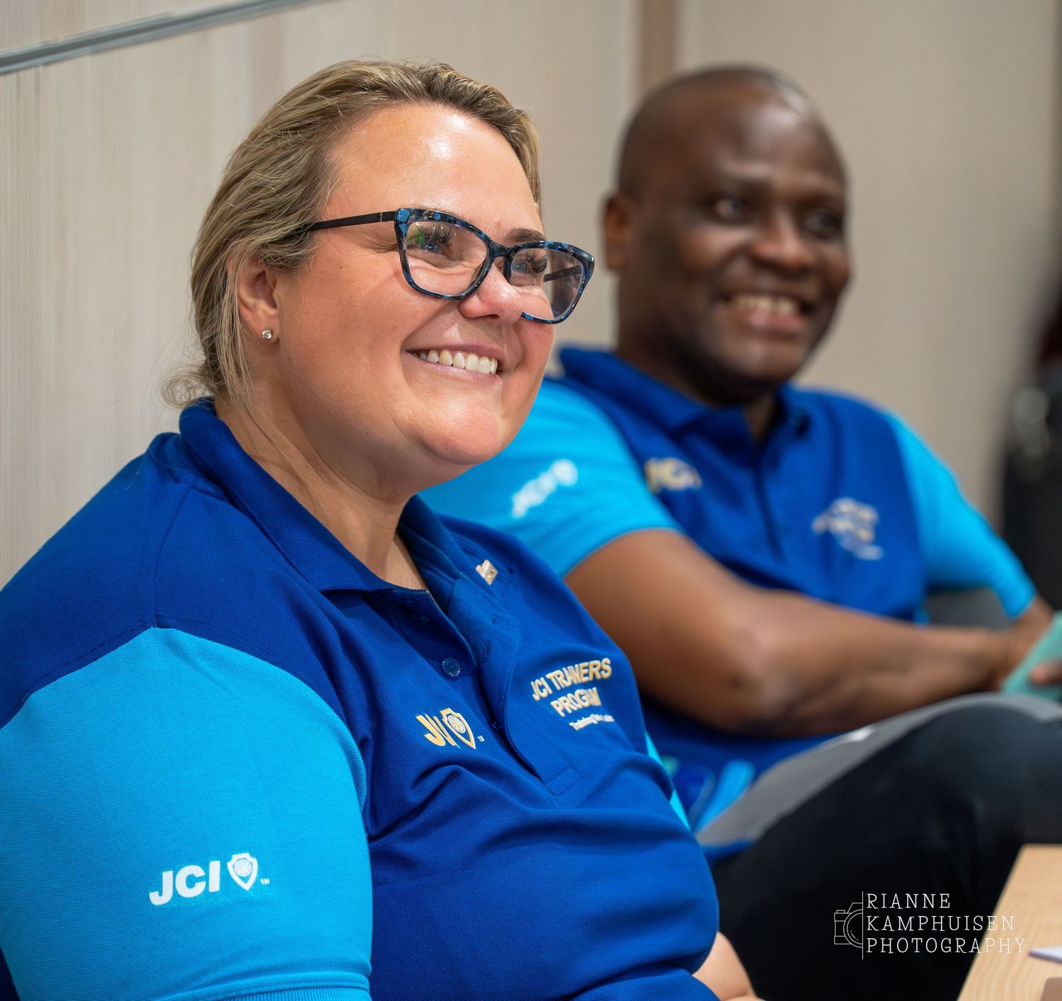 Twee deelnemers in blauwe JCI-shirts lachen tijdens een bijeenkomst, zittend naast elkaar.