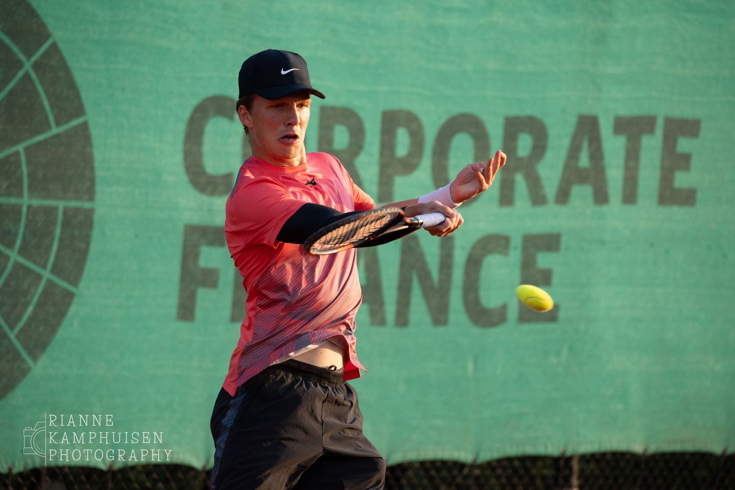 Speler in rood shirt en zwarte pet slaat een krachtige forehand op een gravelbaan.