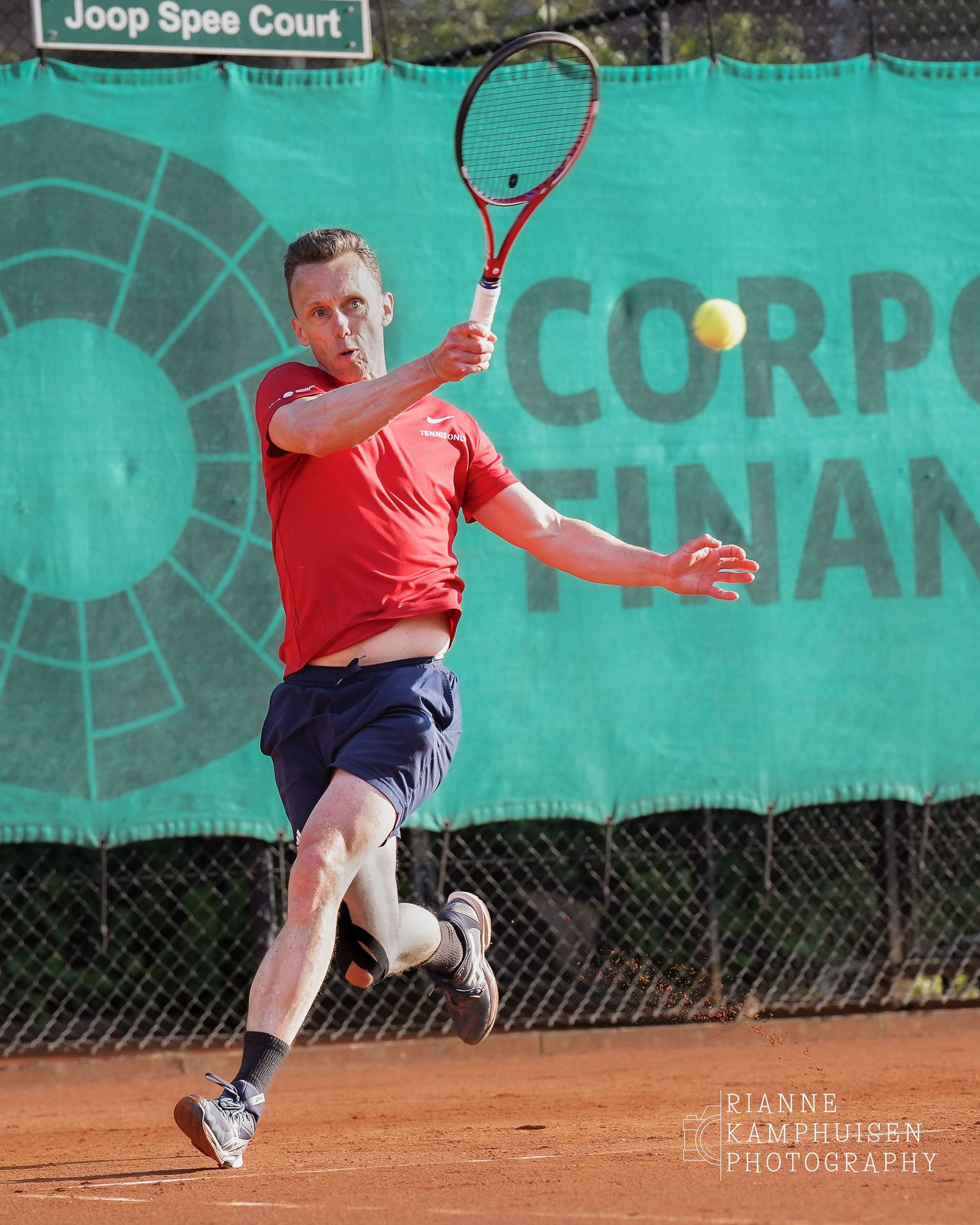 Speler in rood shirt maakt een krachtige forehand op het gravel tijdens een wedstrijd.