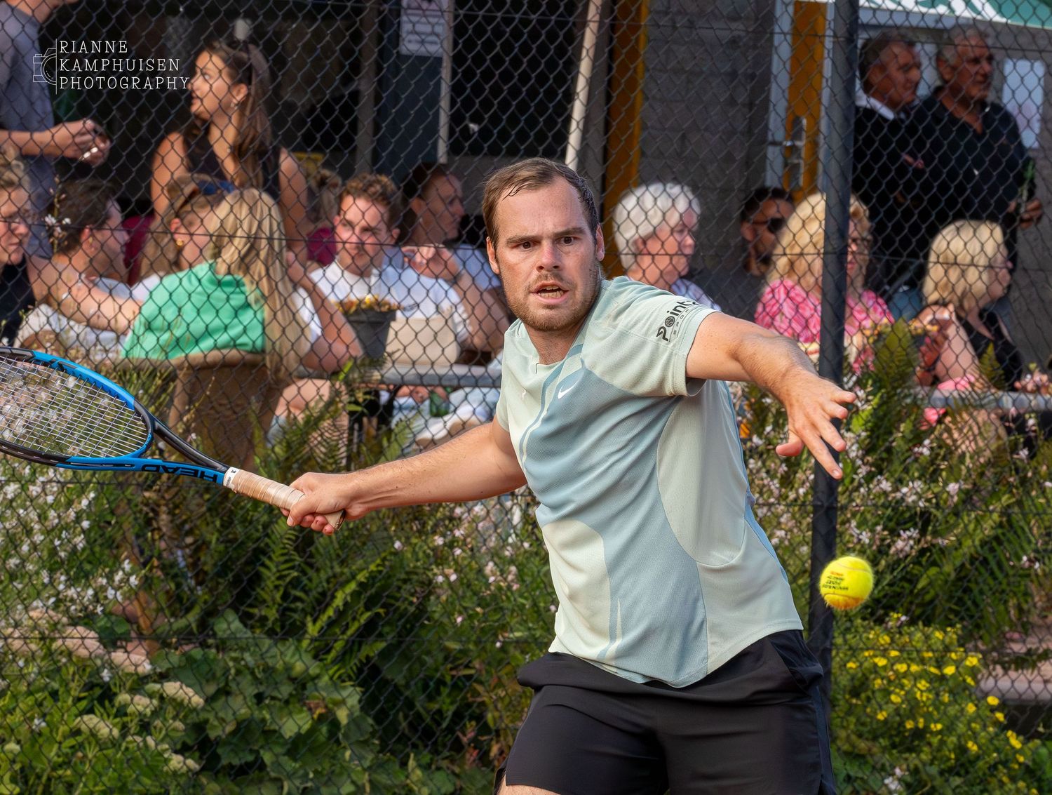 Tennisser in actie slaat een forehand, met toeschouwers op de achtergrond achter een hek.
