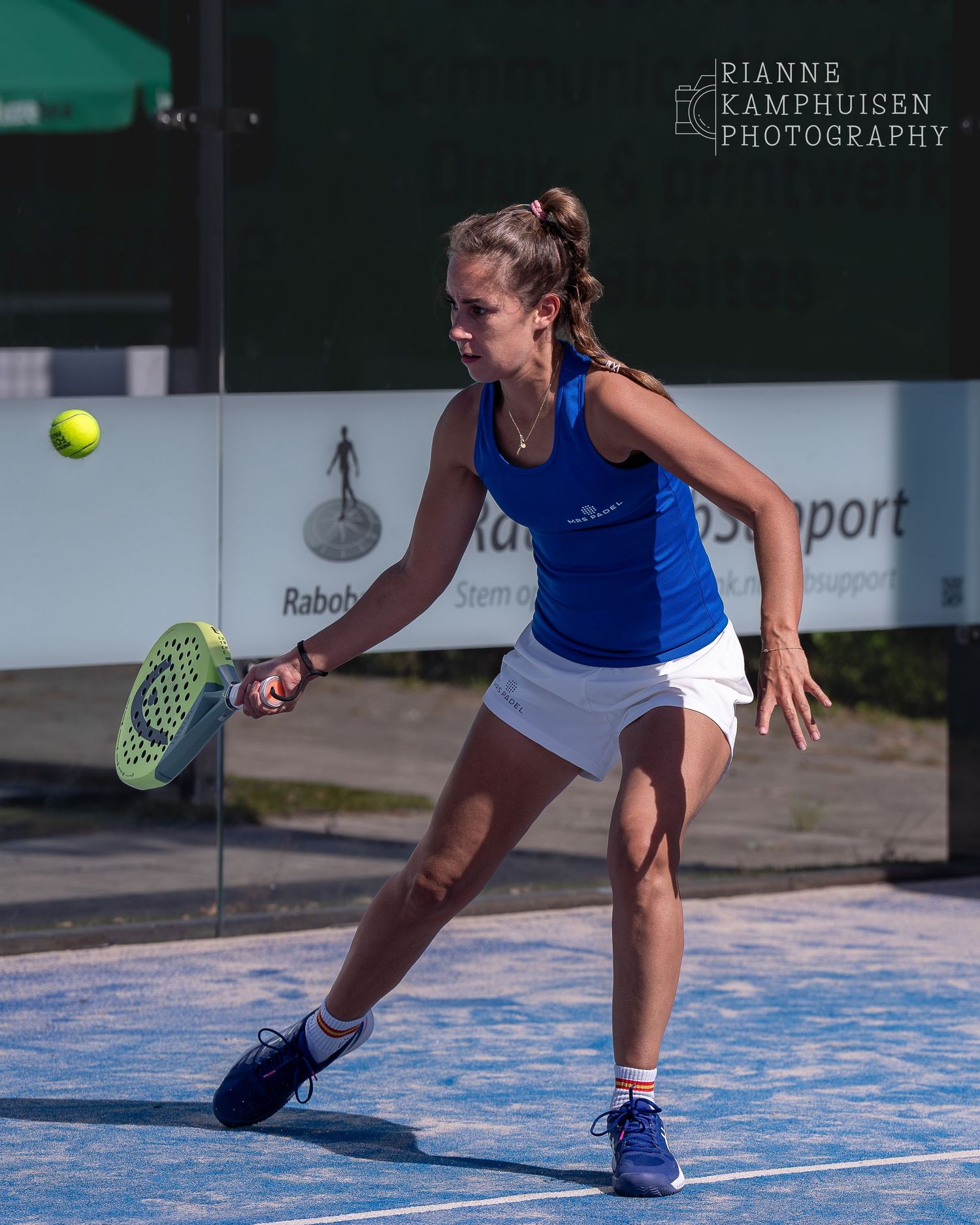 Jonge vrouw in blauw topje en wit rokje slaat een forehand op de zadelbaan