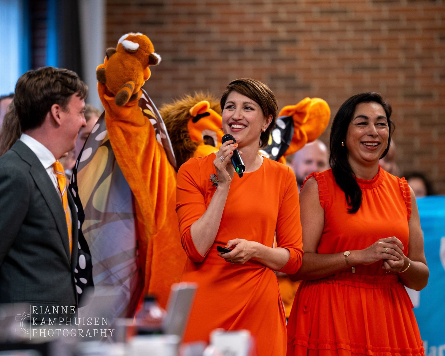 Twee vrouwen in oranje jurken lachen tijdens een presentatie, met een leeuwenmascotte en andere deelnemers op de achtergrond.