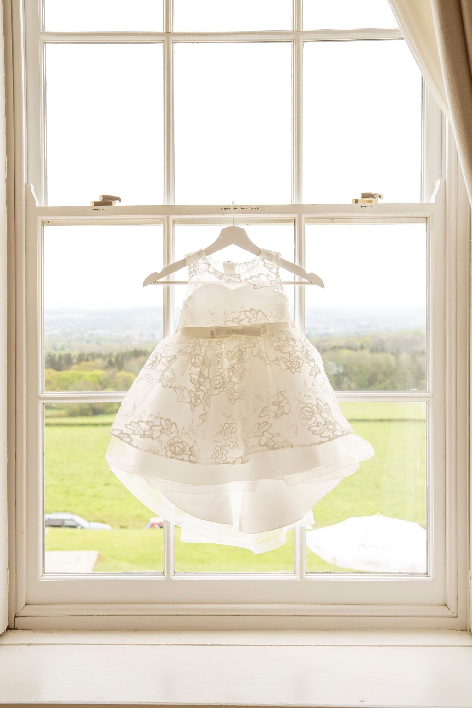 Vintage white wedding dresses hang on wooden hangers against sunlit windows overlooking green countryside meadows.