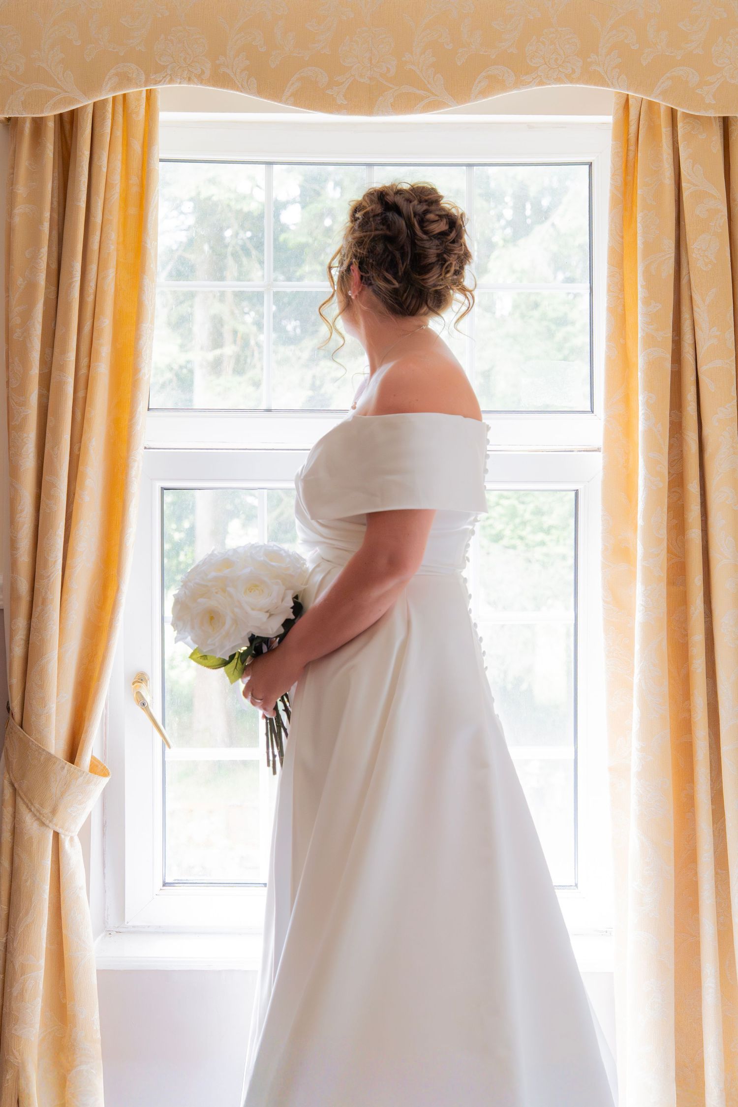 Bride in white off-shoulder gown holding bouquet, standing by sunlit window with golden curtains.