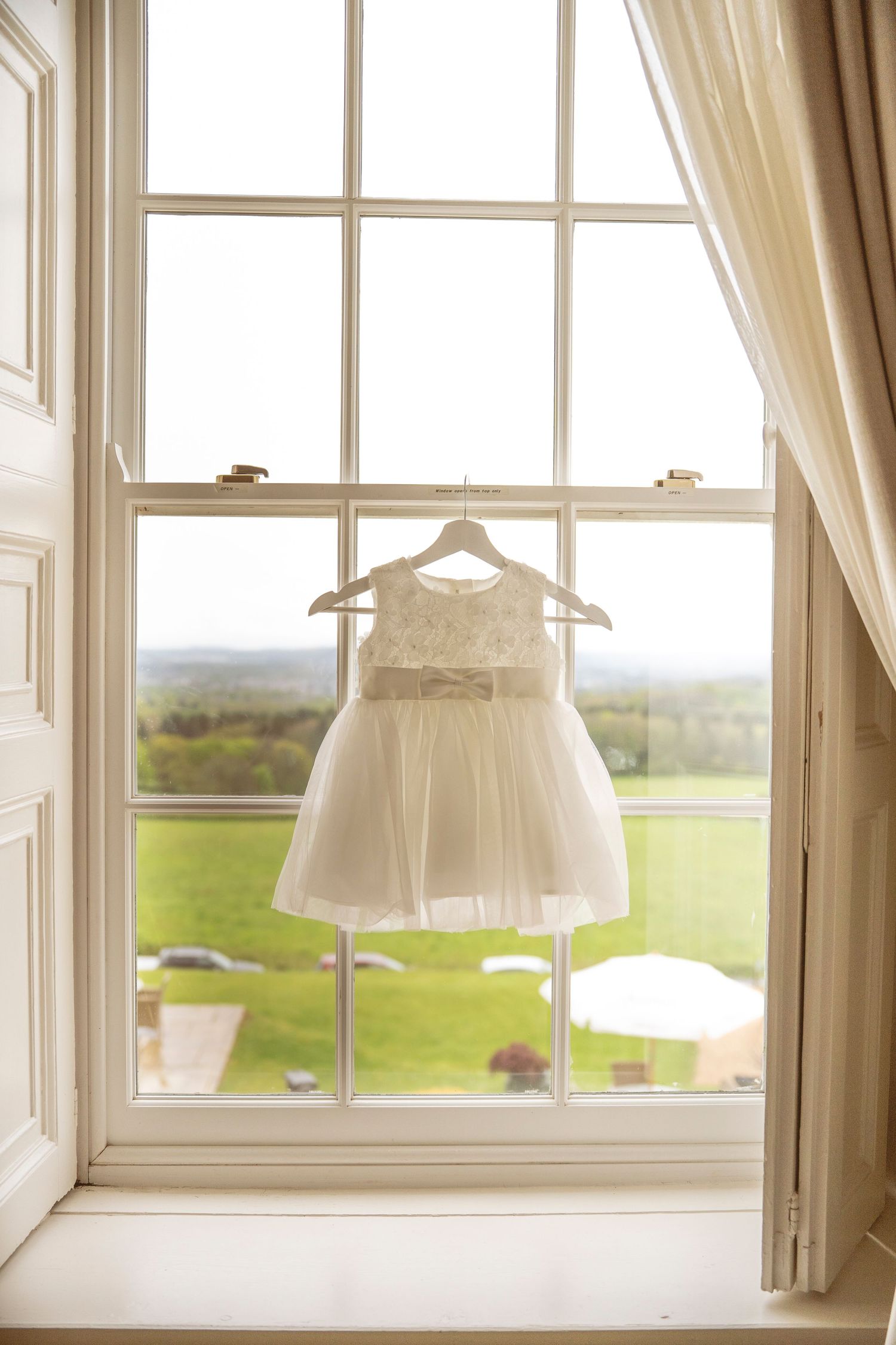 White baby dresses hanging by windows overlooking a scenic green landscape.