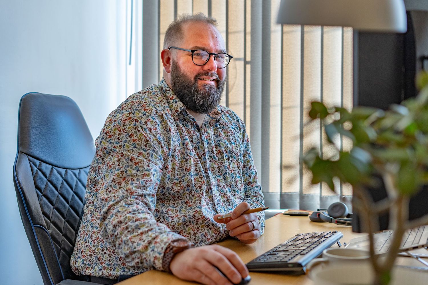 Cheerful bearded professional working at desk with laptop in bright modern office setting with plants.