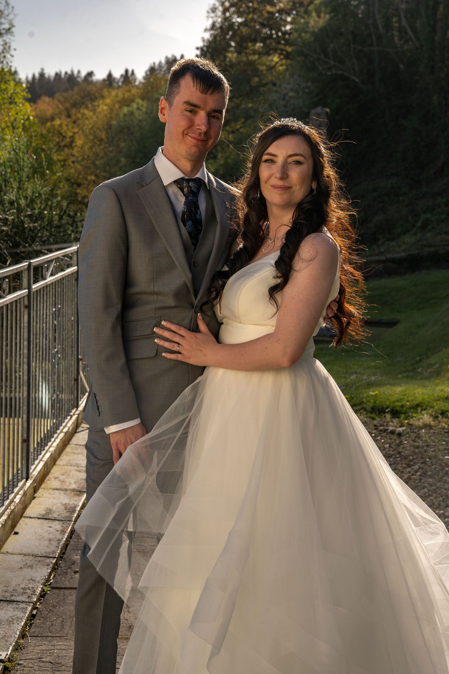 Newlywed couple posing on outdoor bridge with autumn foliage in background.