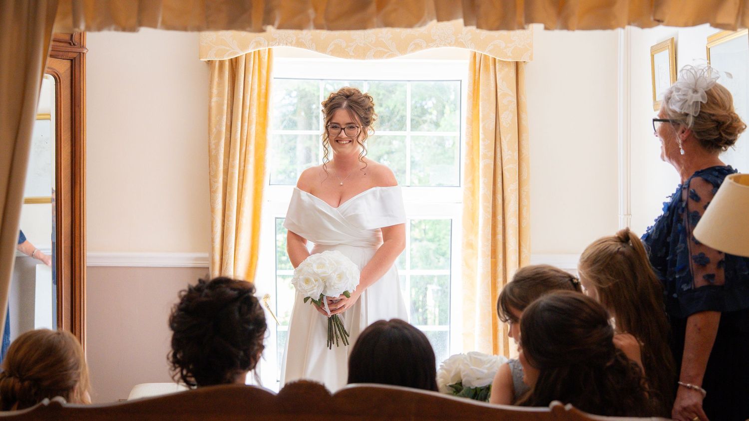 Bride in white off-shoulder gown holding bouquet stands before window at indoor wedding ceremony with seated guests.