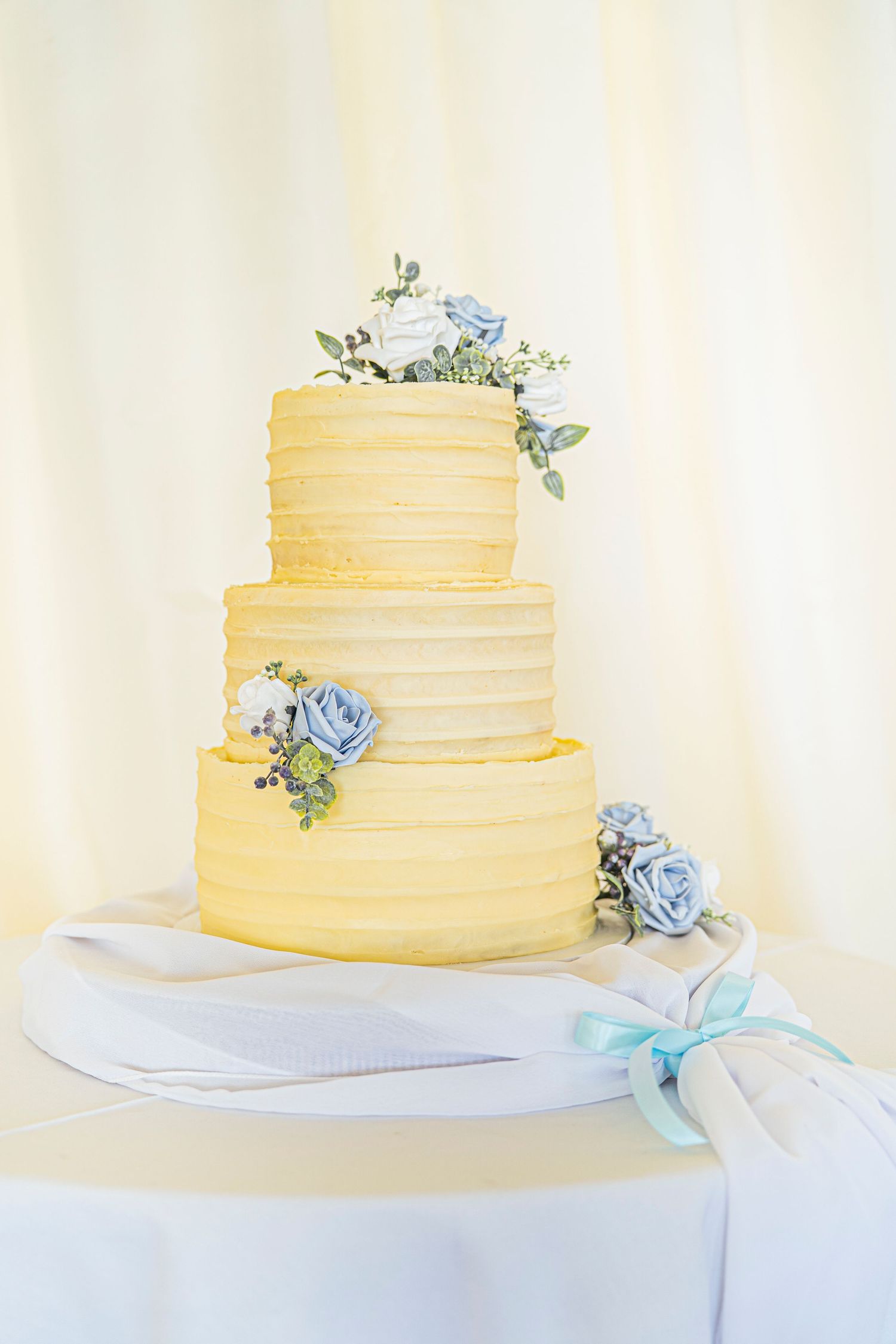 Three-tiered buttercream wedding cake with delicate blue roses and greenery decorations on a white stand.