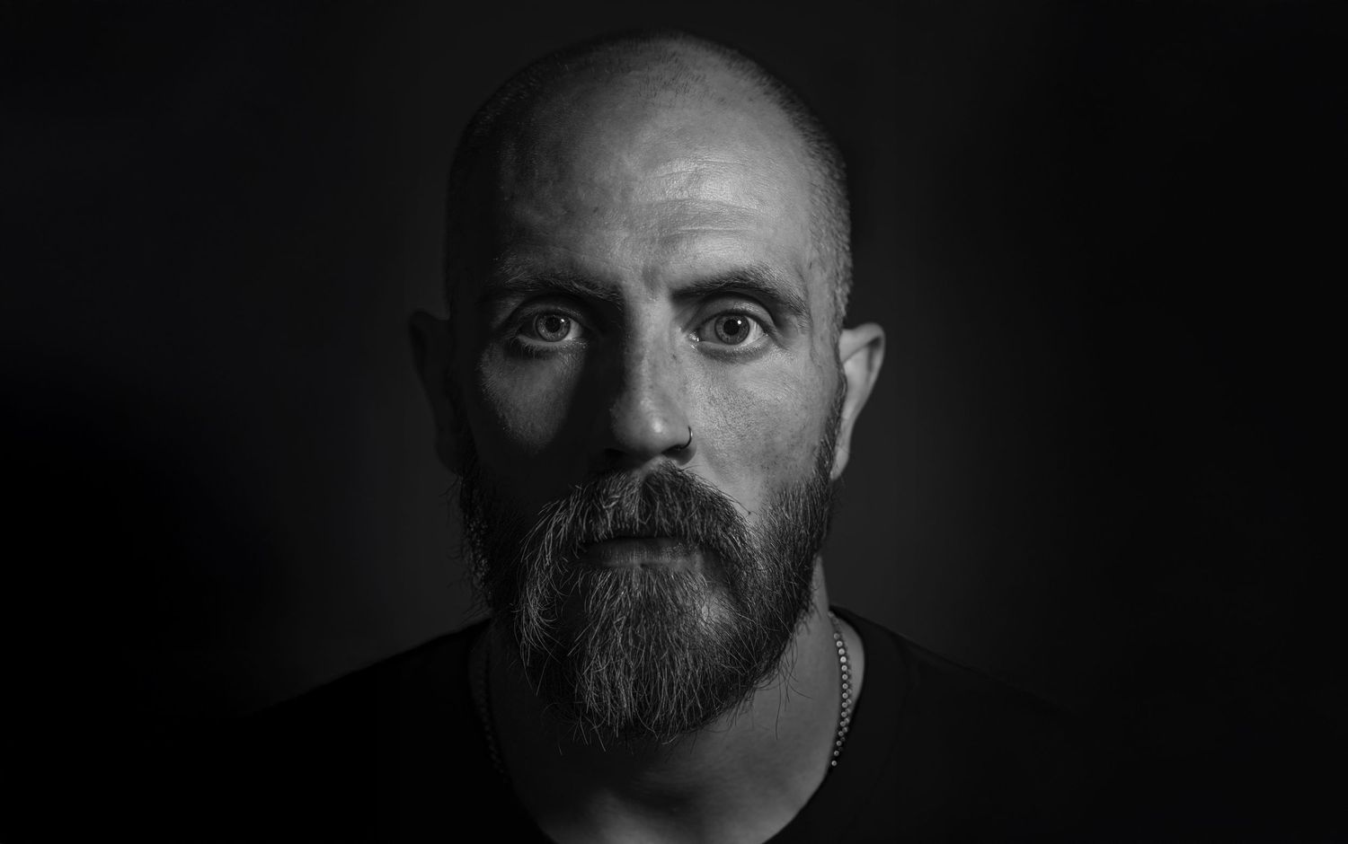 Dramatic black and white portrait of a person with a full beard against dark background.