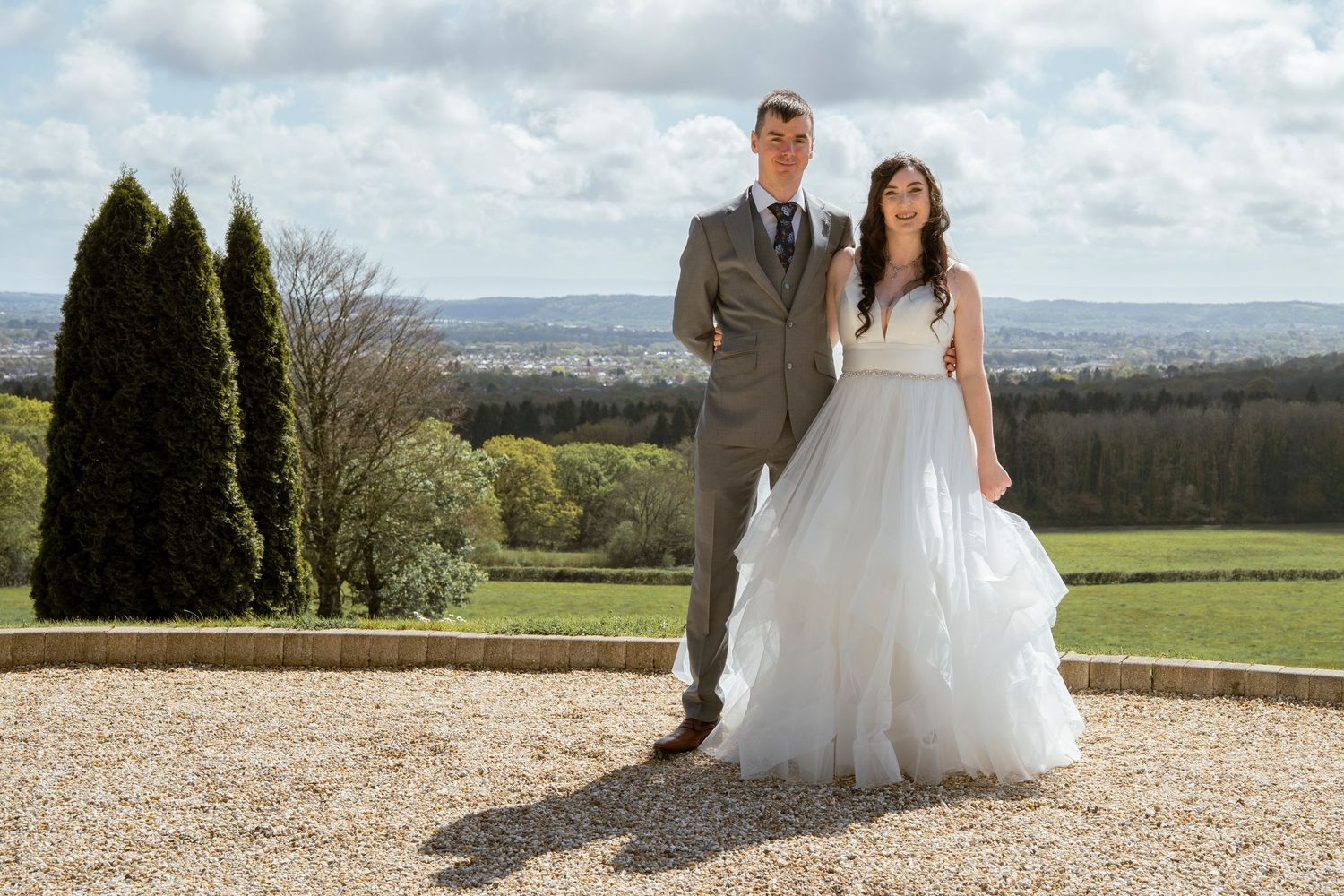 Panoramic countryside view forms backdrop for wedding photos on gravel path.