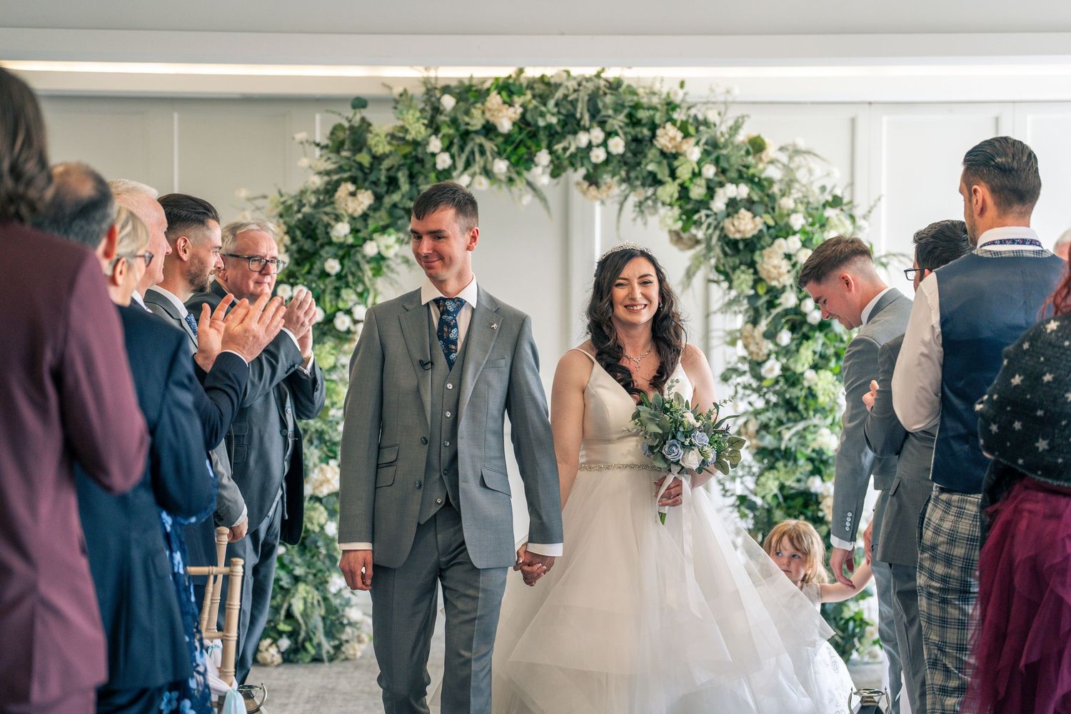 Wedding ceremony scene with floral arch decorations and guests gathered in an indoor venue.