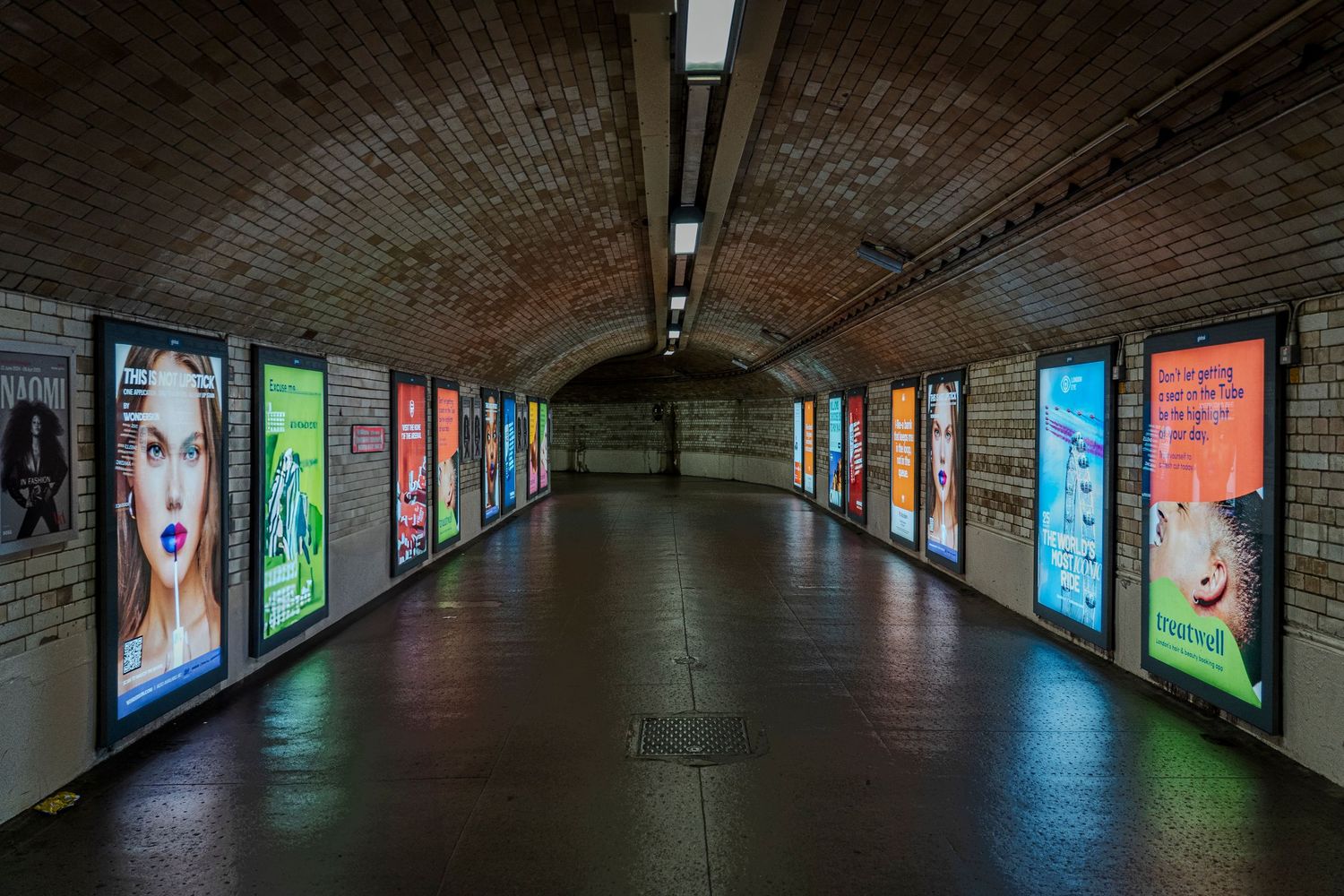 A long underground tunnel with brick walls and illuminated advertising displays on both sides.