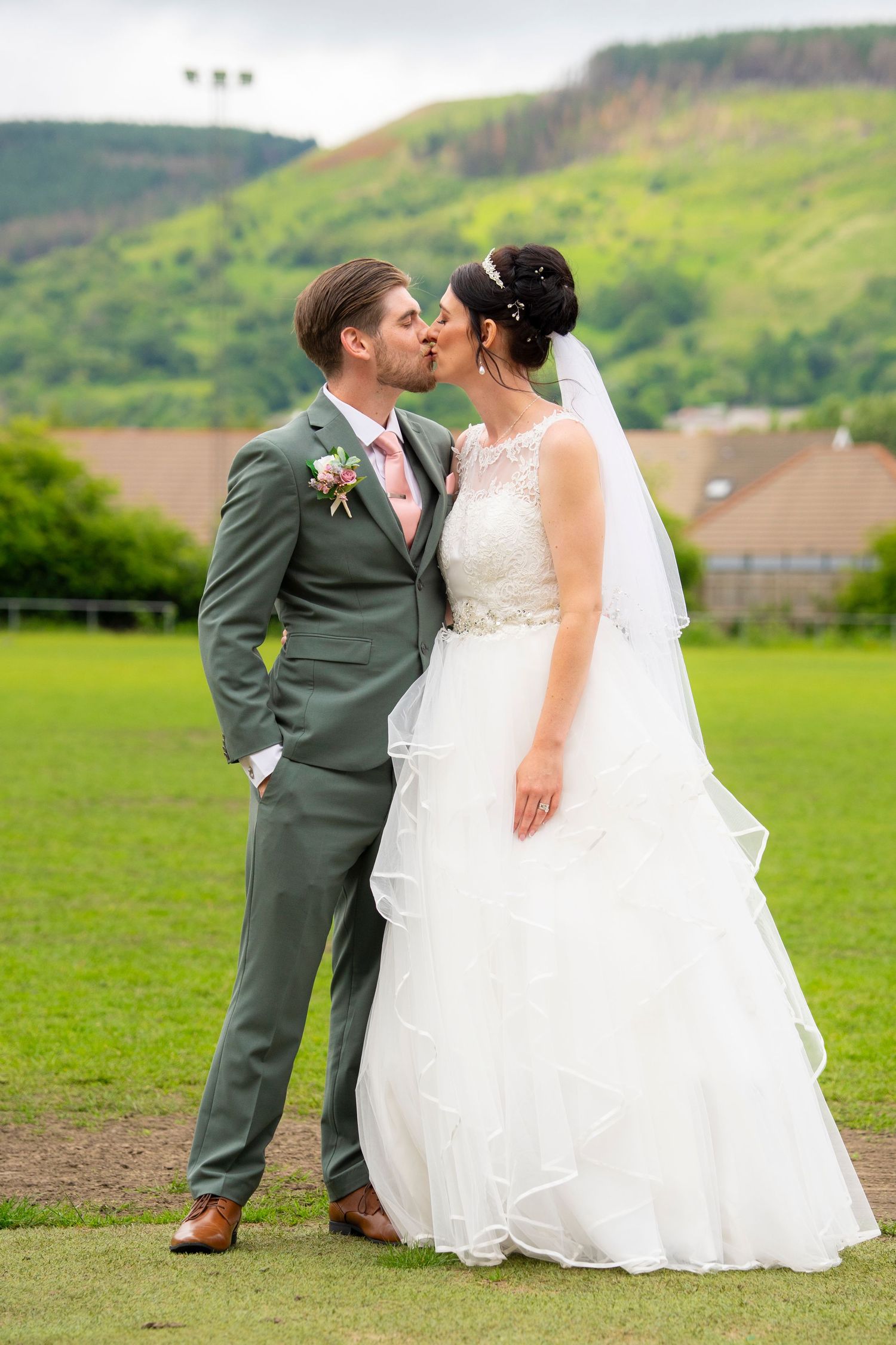 Romantic newlyweds share tender moments in a green countryside field, featuring white wedding dress and gray suit attire.