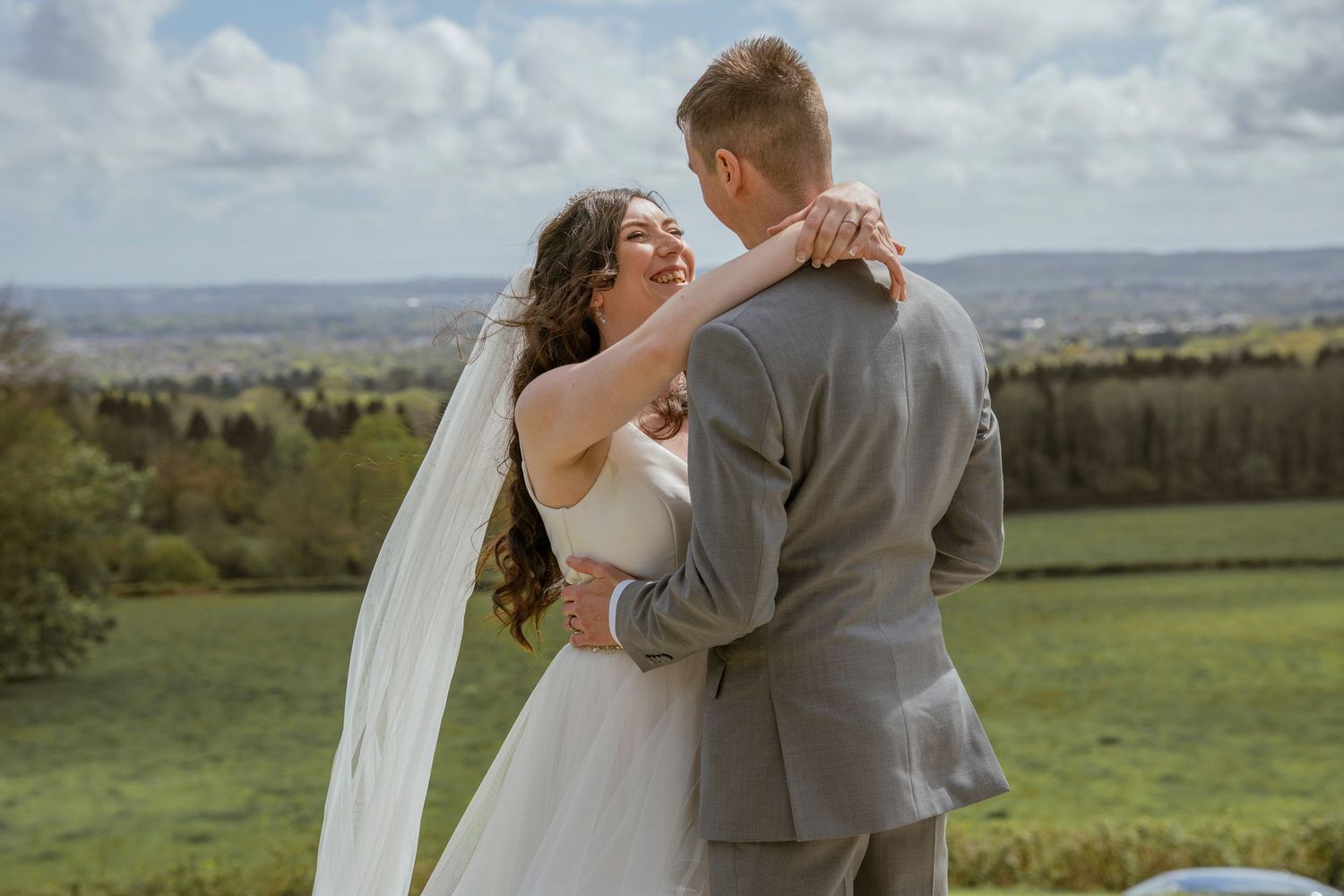 Couple embracing in wedding attire on a hilltop overlooking a scenic landscape.
