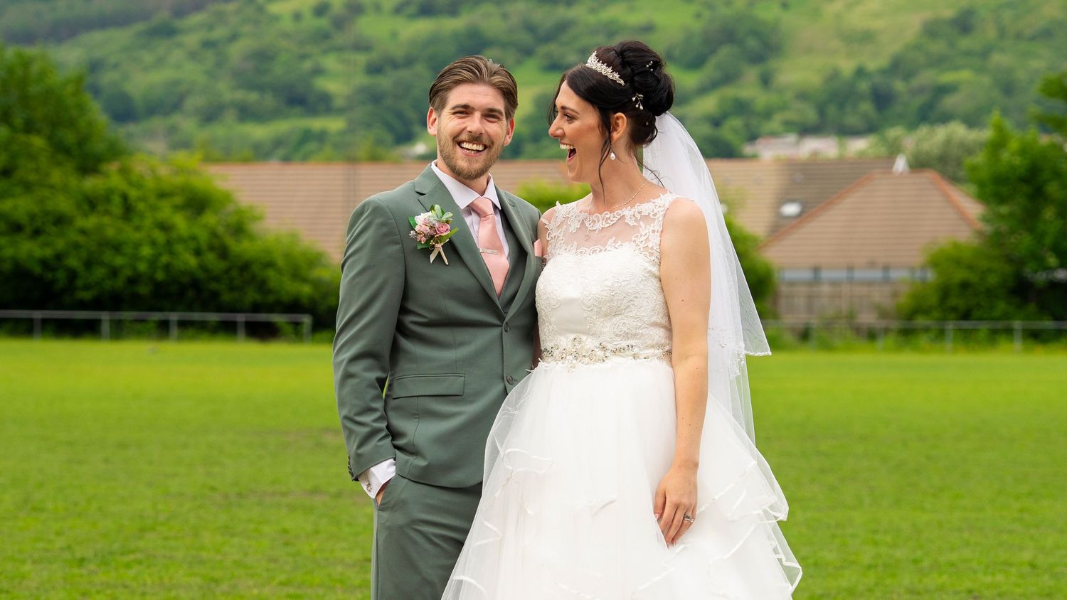 Wedding couple posing in a lush green field with mountains in the background.