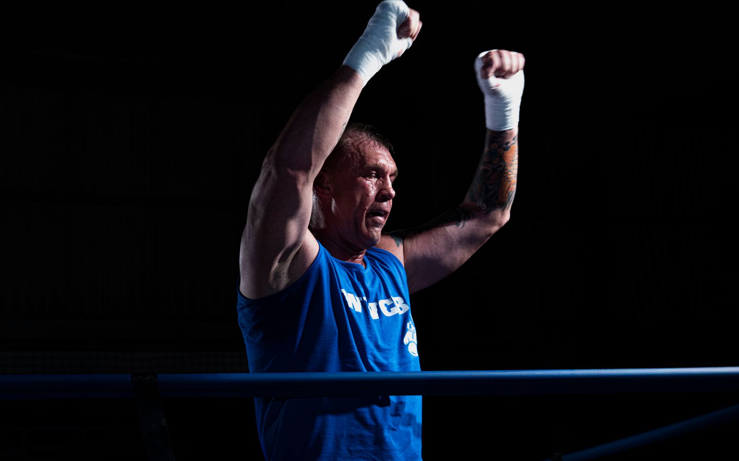 Athlete in blue uniform raises arms in celebration during competitive match against dark background.