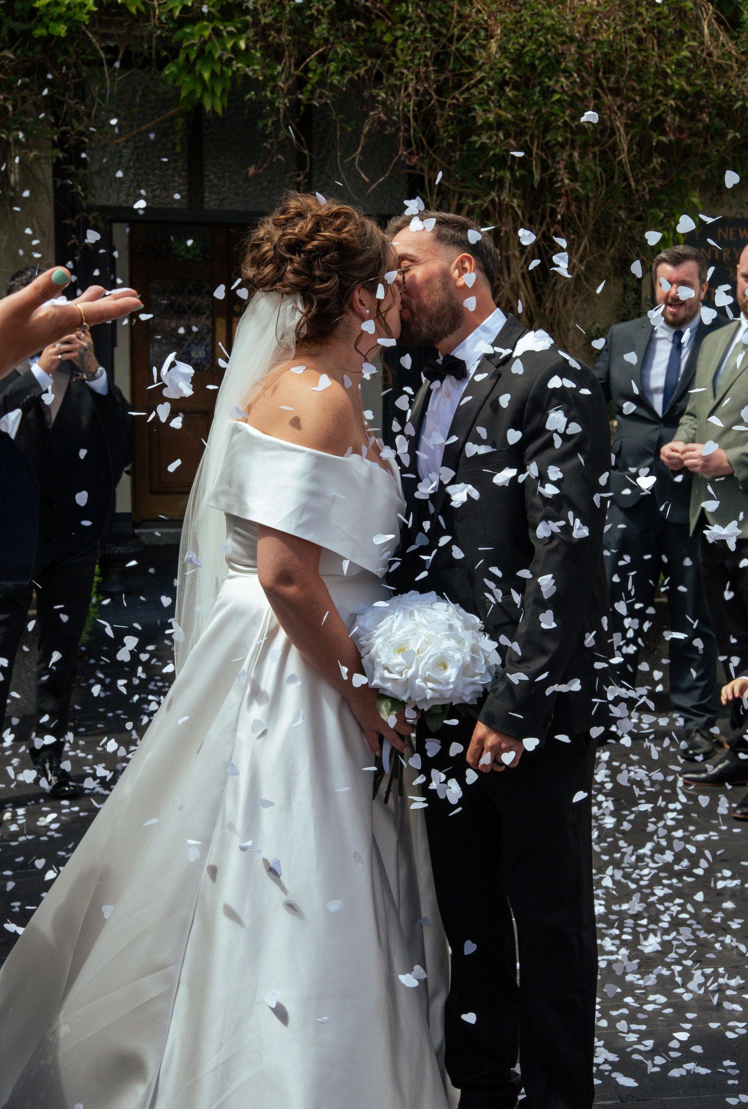 Bride and groom share a kiss surrounded by white confetti hearts during outdoor wedding ceremony.