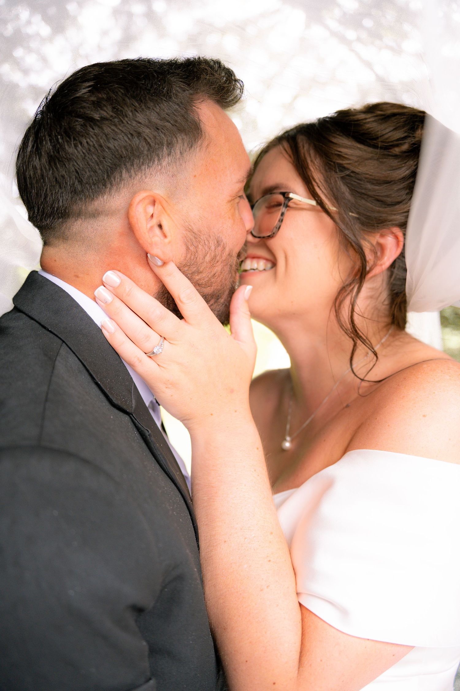 Bride and groom sharing a romantic moment under a veil, smiling closely with wedding ring visible on her hand.