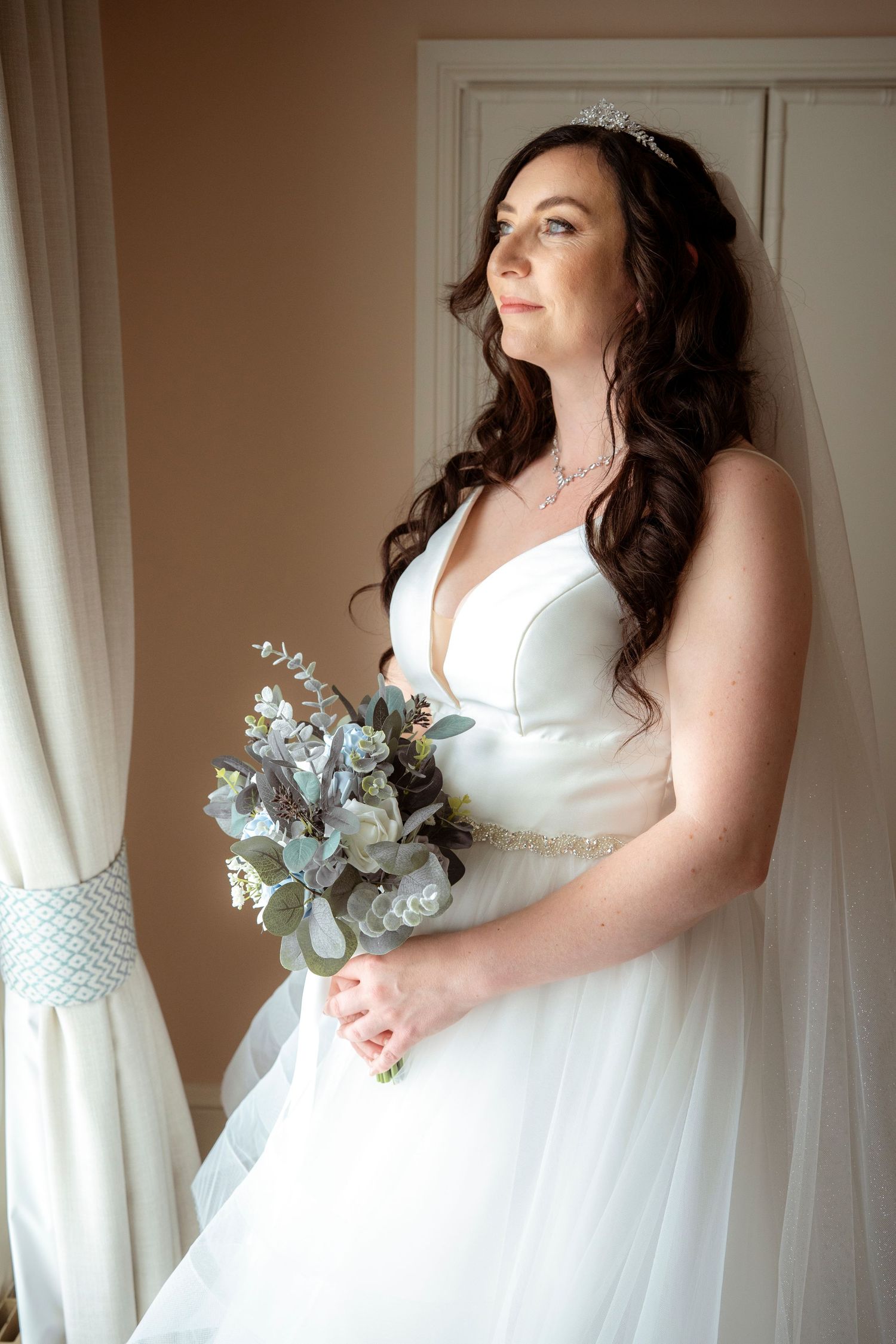 A bride in a white wedding dress holds a delicate bouquet with blue-gray succulents near a window.