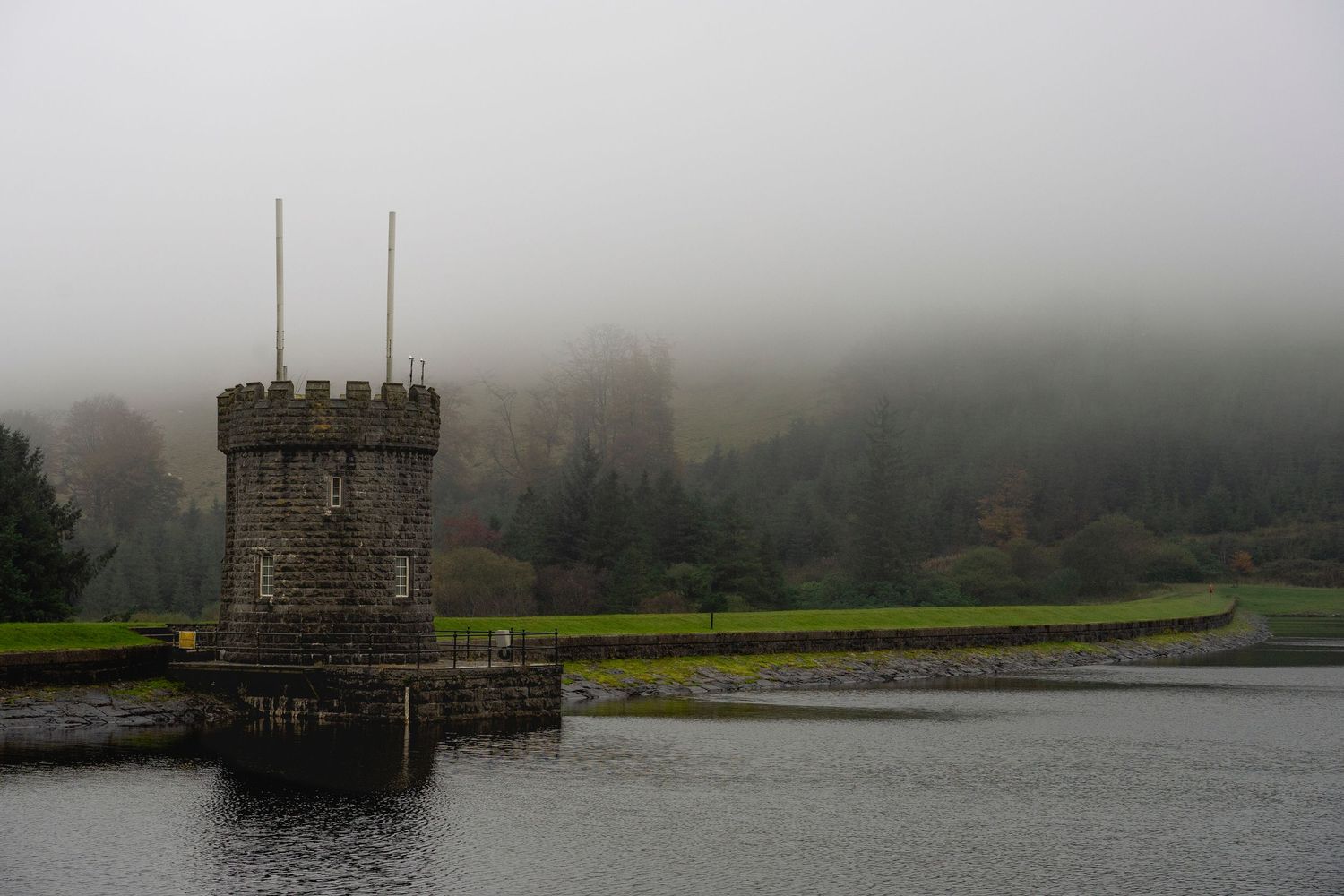 An old stone tower stands in misty weather beside a reservoir in the countryside.