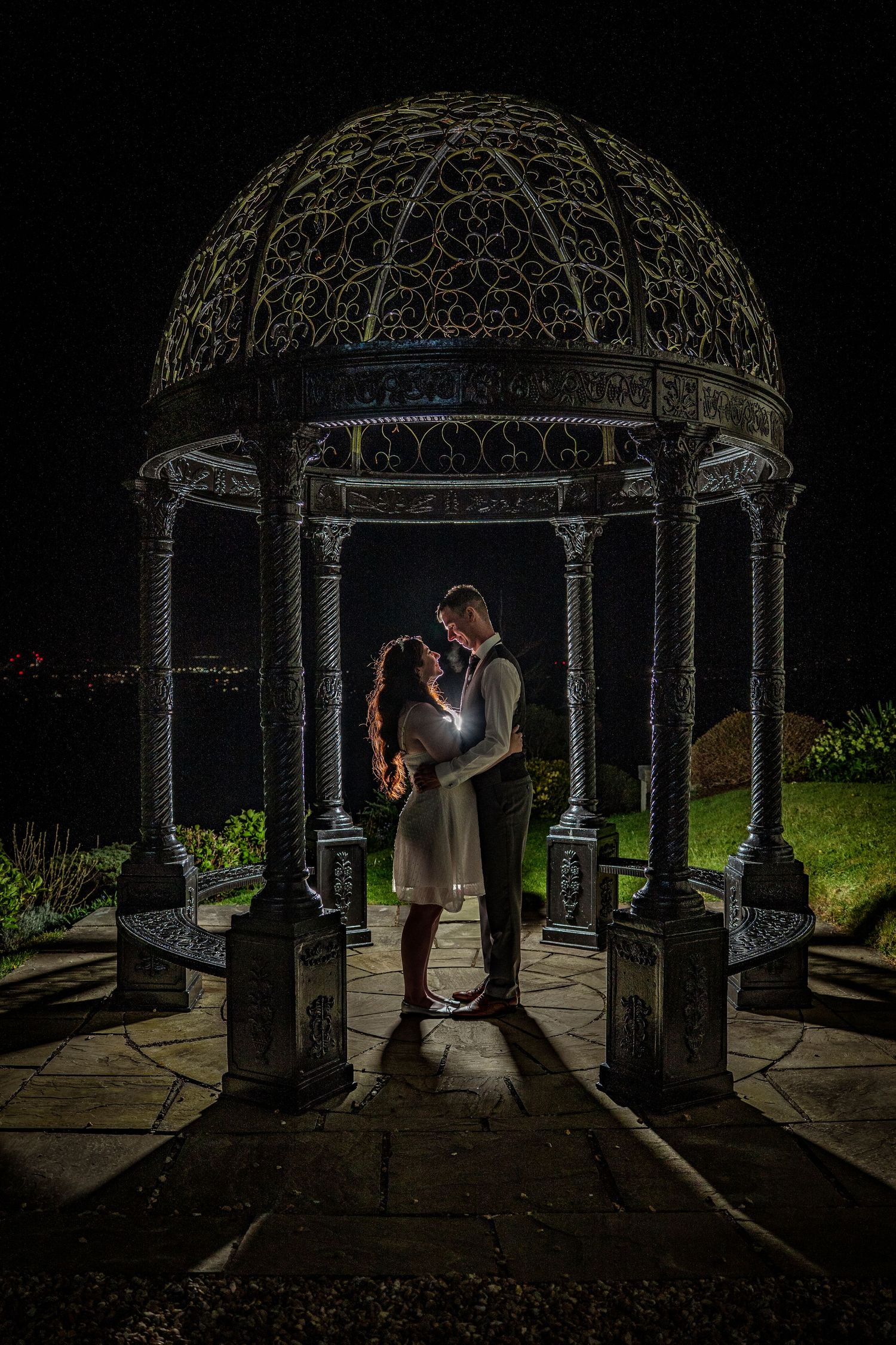 Silhouetted couple shares intimate moment under illuminated garden gazebo at night.