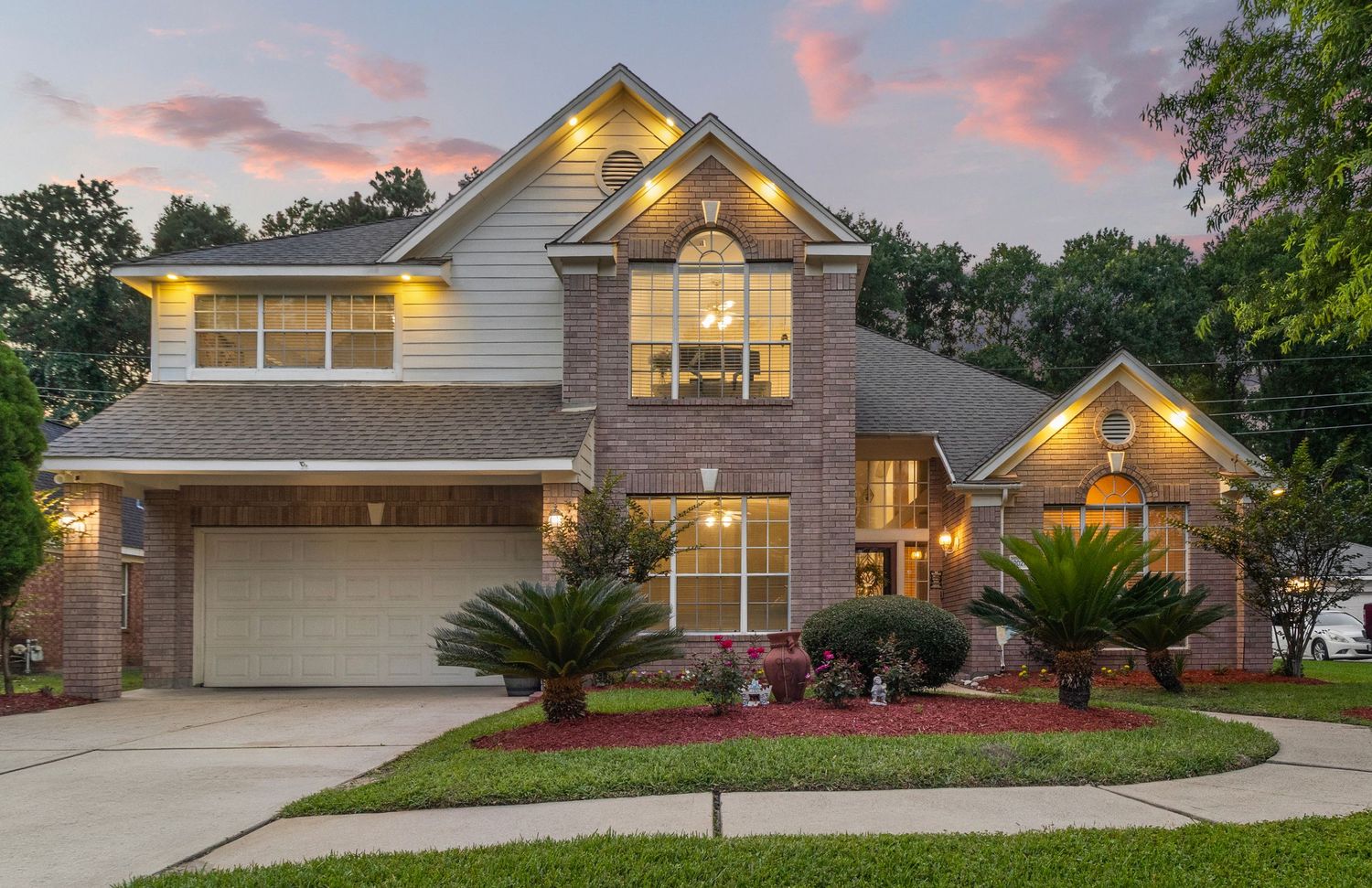 Two-story brick home with glowing windows at dusk, manicured landscaping, and a two-car garage in a suburban neighborhood.