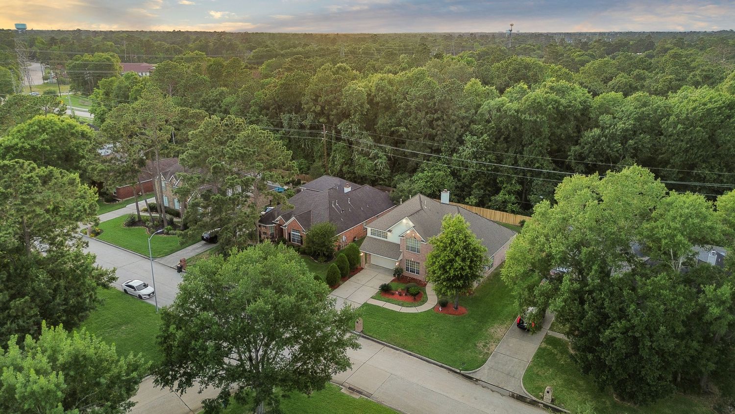 Aerial view of a large brick home surrounded by lush green trees in a serene suburban neighborhood at dusk.