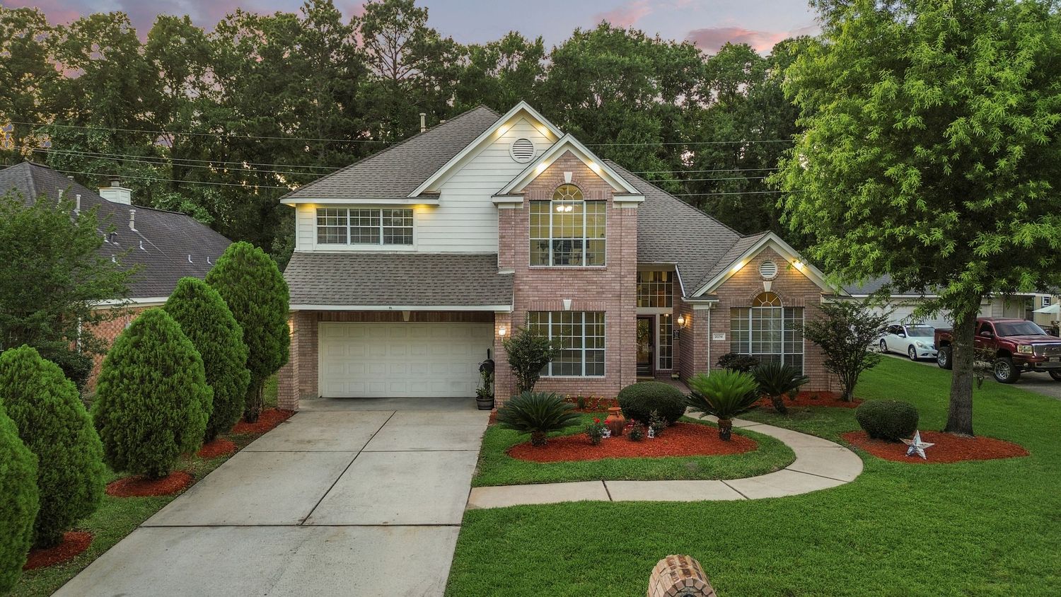 Two-story brick home with manicured lawn, lush trees, concrete driveway, and warm exterior lighting at dusk.