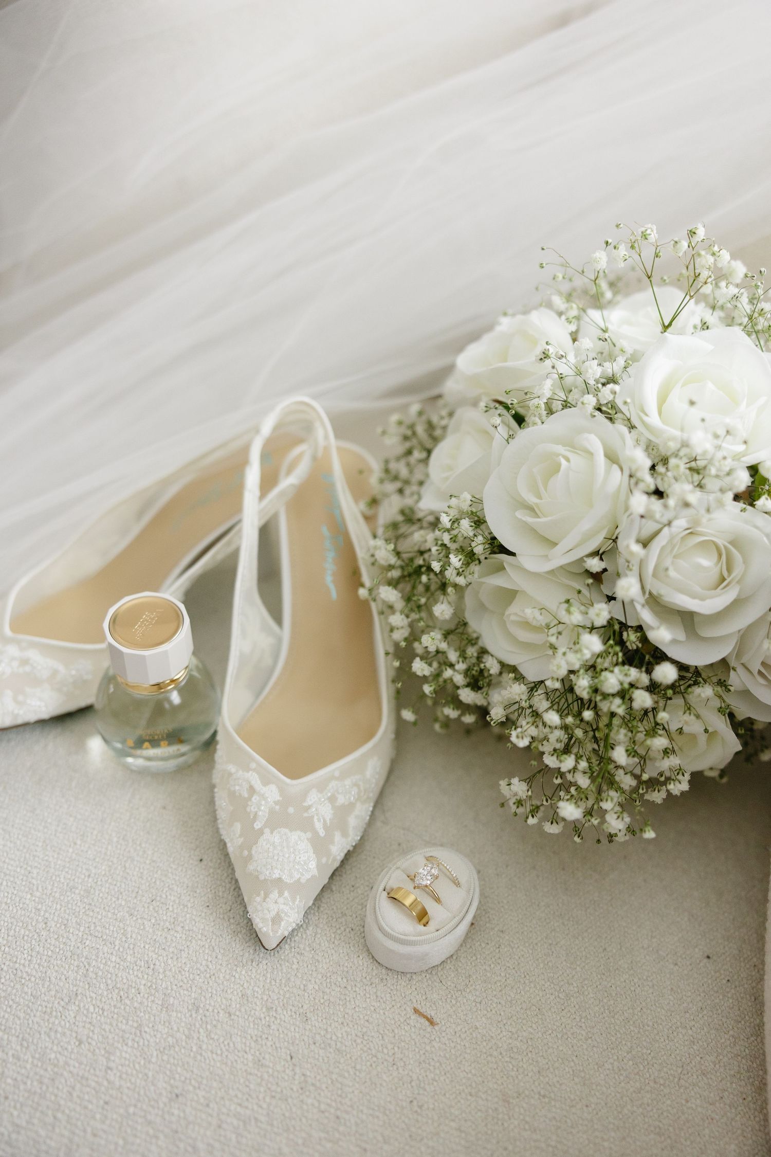White lace bridal heels beside a bouquet of white roses, gold rings in a box, and perfume on a soft white surface.