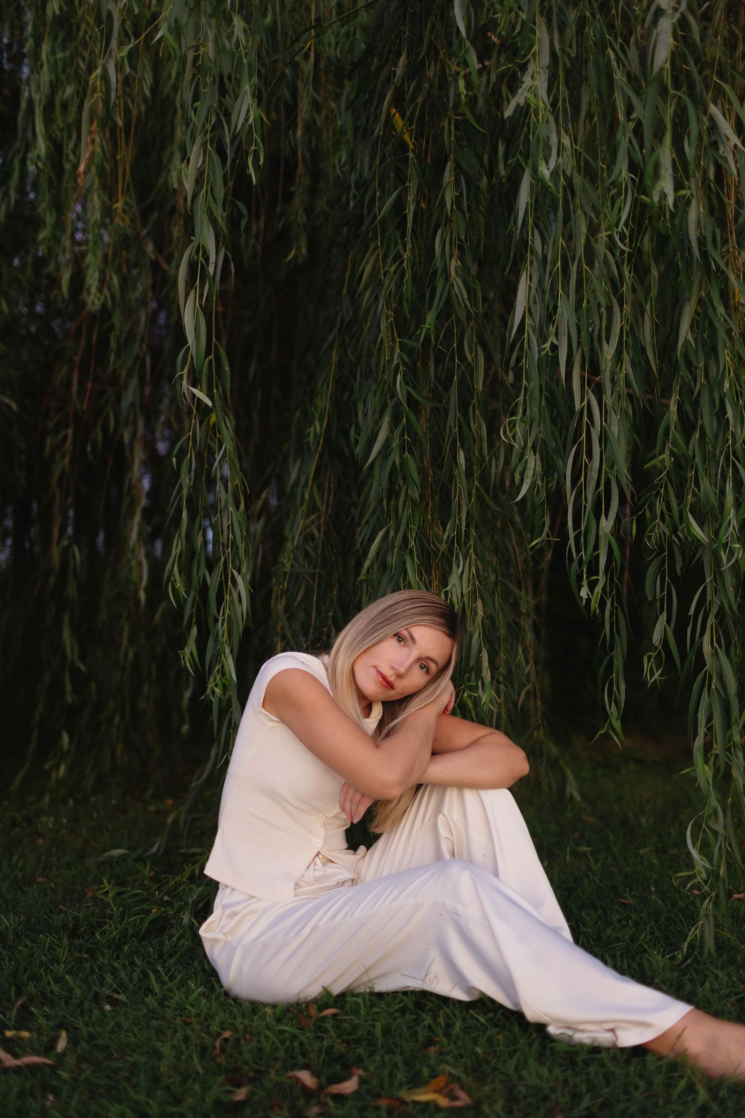 Woman in white outfit sitting peacefully on grass beneath drooping weeping willow tree branches.