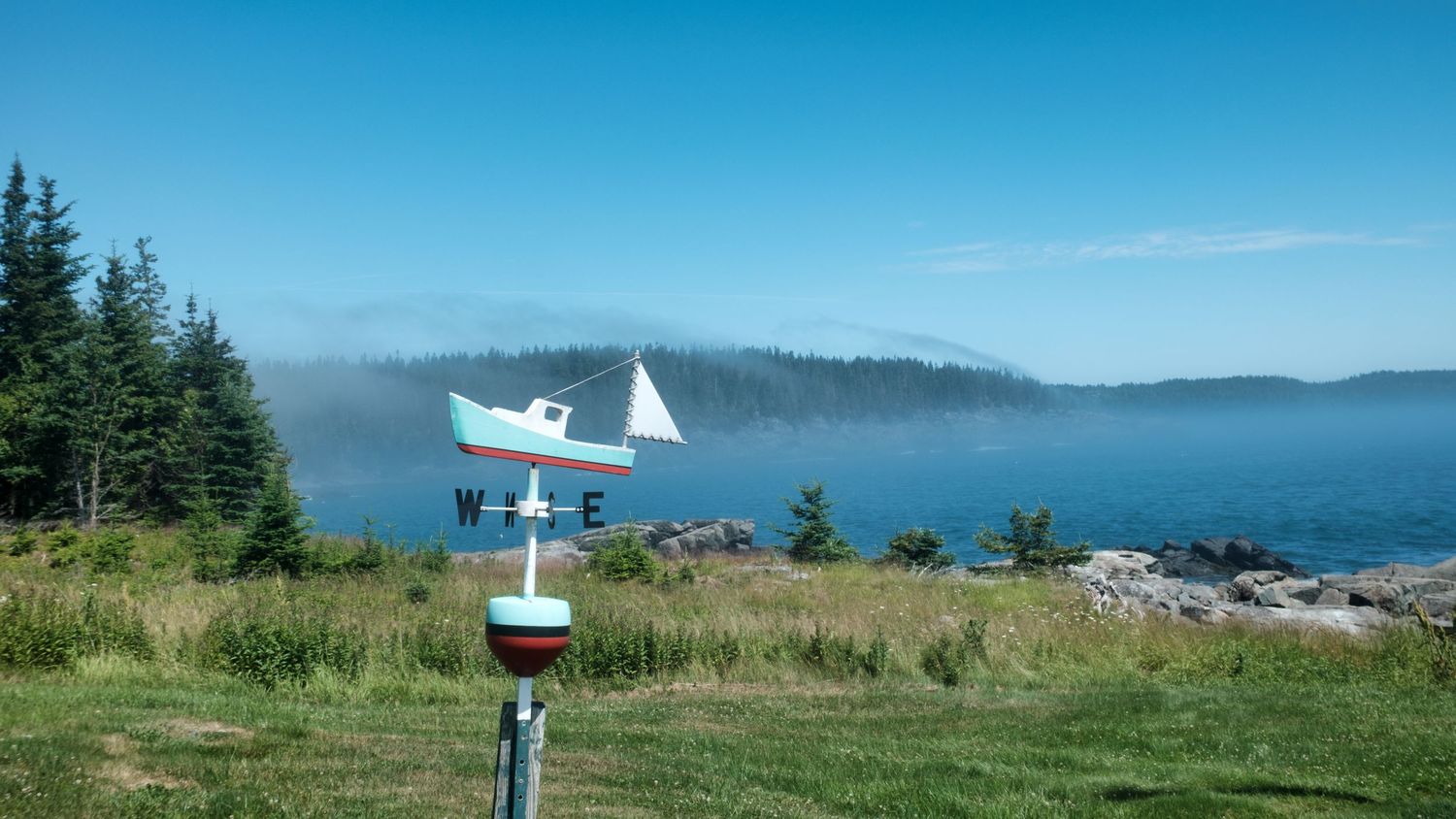 Lighthouse stands on a small island off the coast near Cutler, Maine, surrounded by blue skies and drifting sea fog.