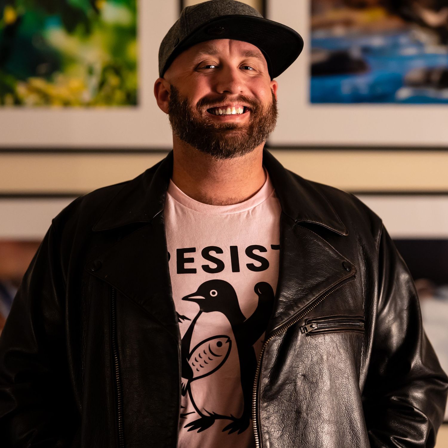 Portrait of photographer Shawn Thomas, smiling in a leather jacket and cap, standing in front of framed prints.
