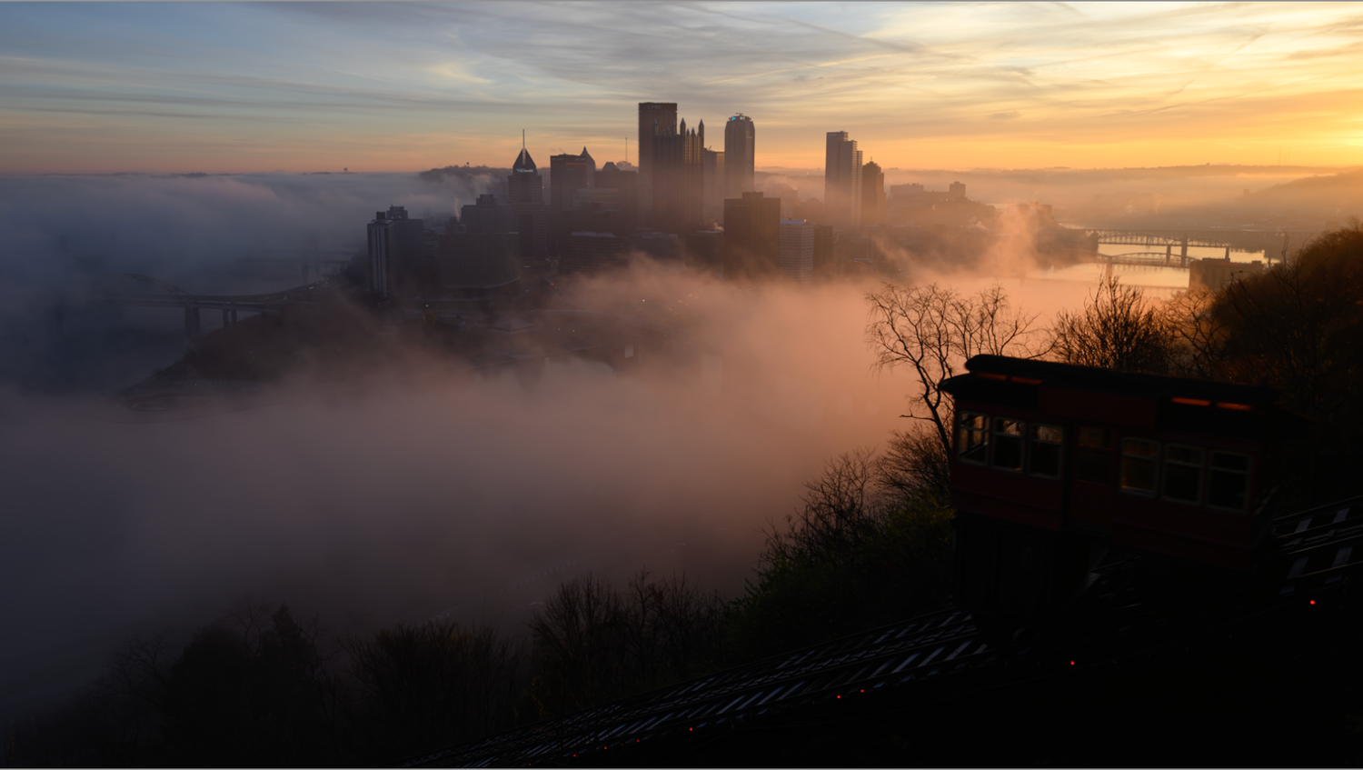Unedited Pittsburgh skyline rises through morning fog as the Duquesne Incline climbs the hillside at sunrise.