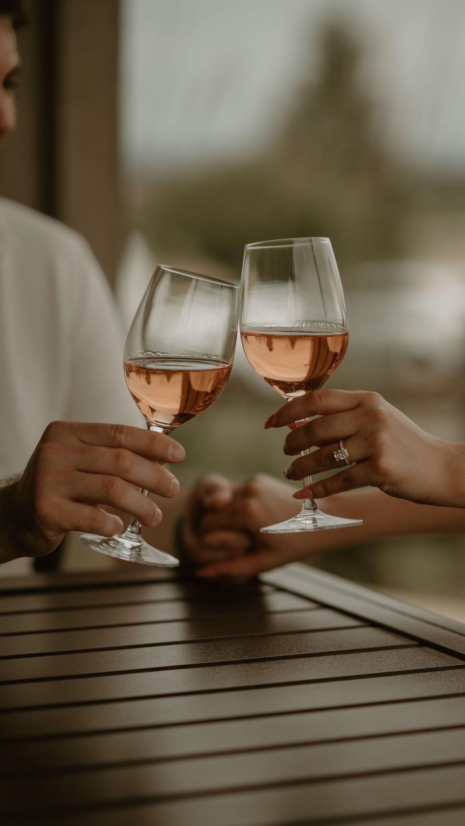 Two hands toasting with glasses of rose wine at sunset on a wooden table outdoors.