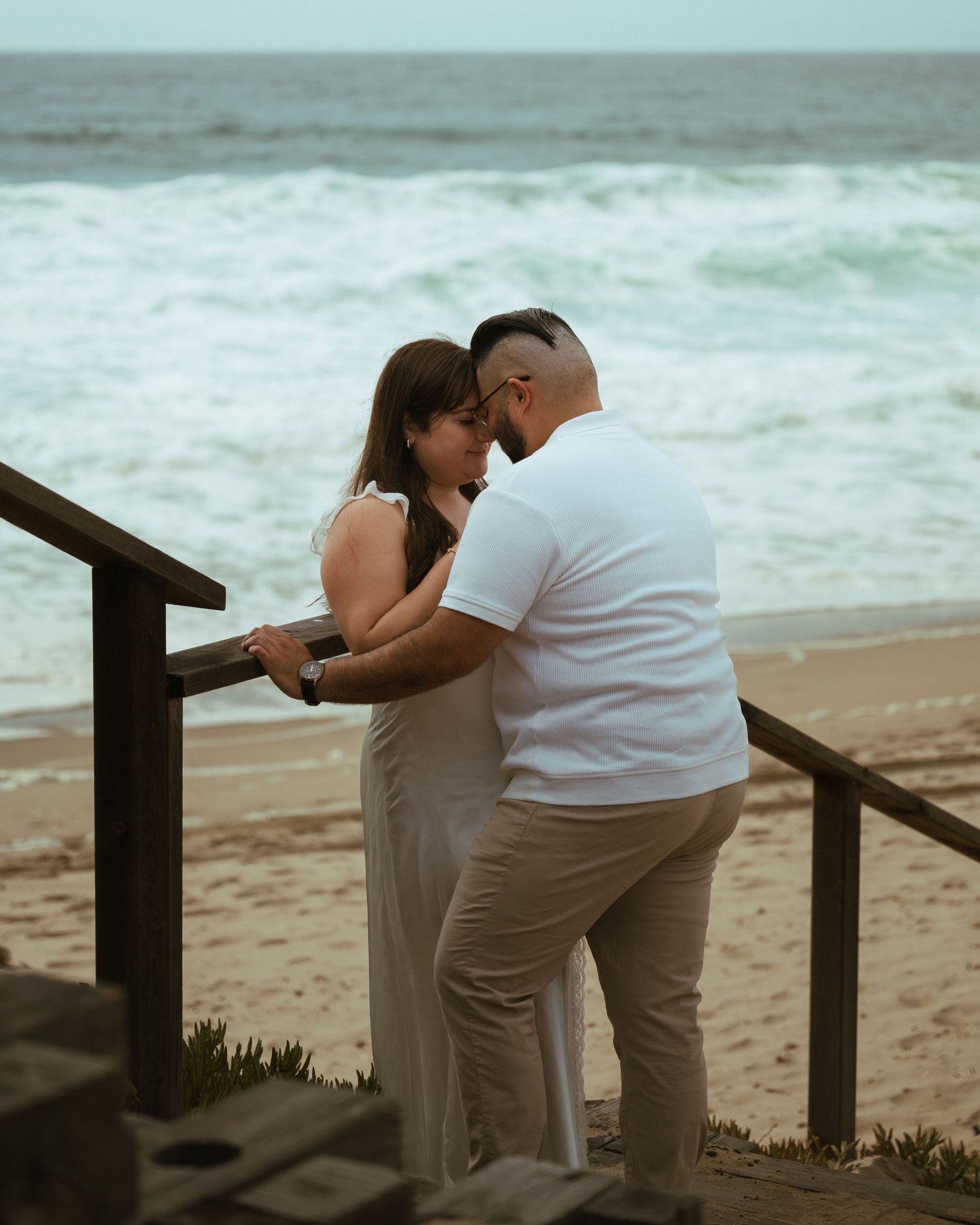 Romantic couple sharing an intimate moment on a beach stairway with ocean waves in the background.