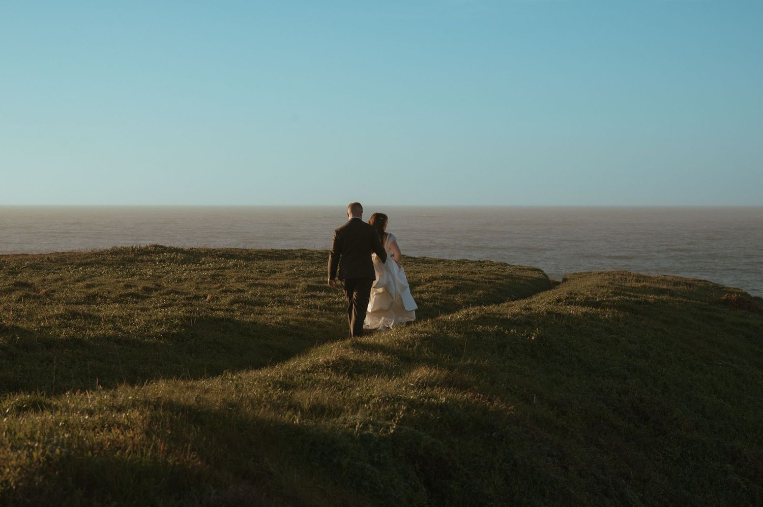 A couple in wedding attire walks hand-in-hand on a grassy coastal cliff overlooking the ocean horizon.
