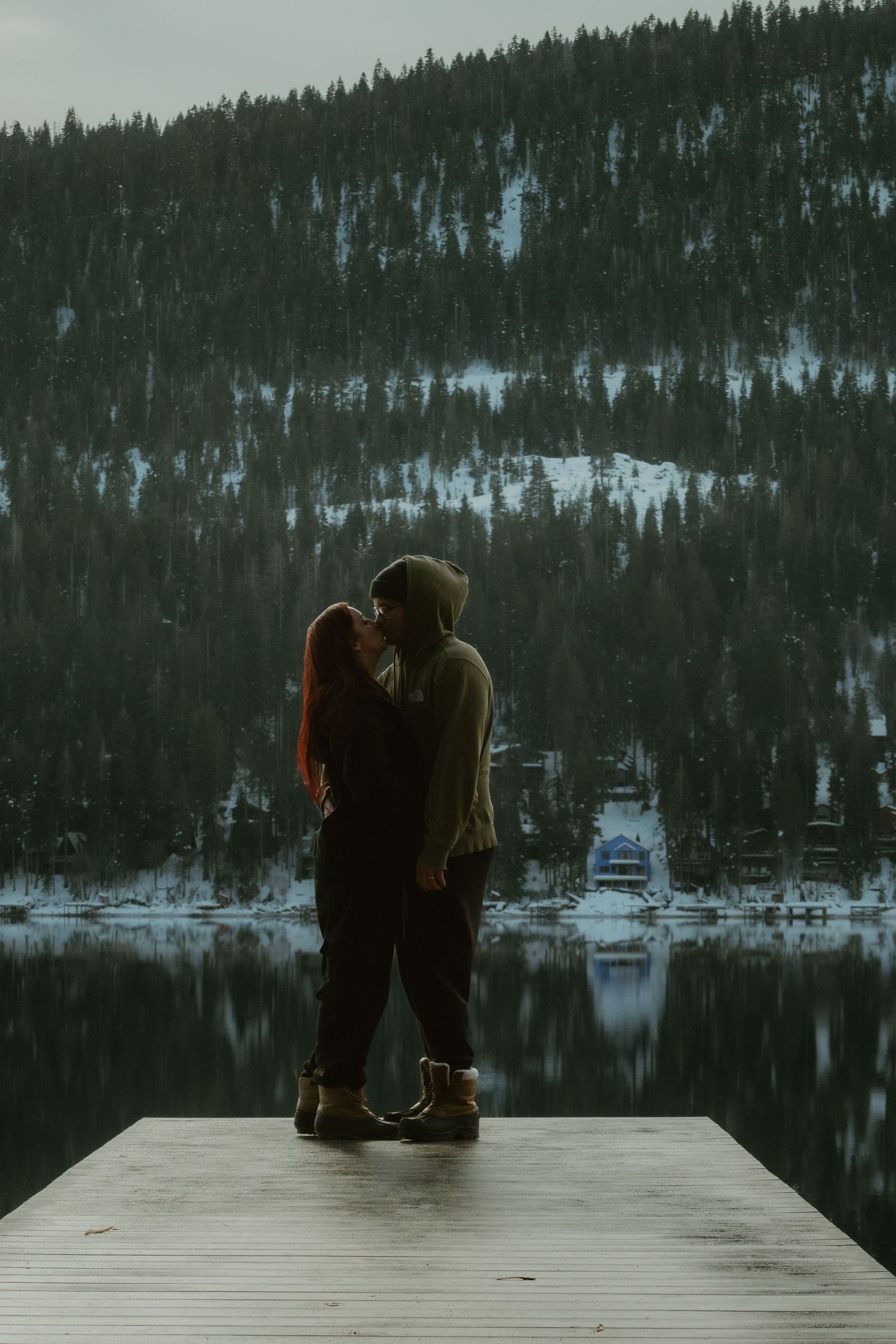 A couple embraces on a wooden dock overlooking a serene lake surrounded by snow-covered mountains at dusk.