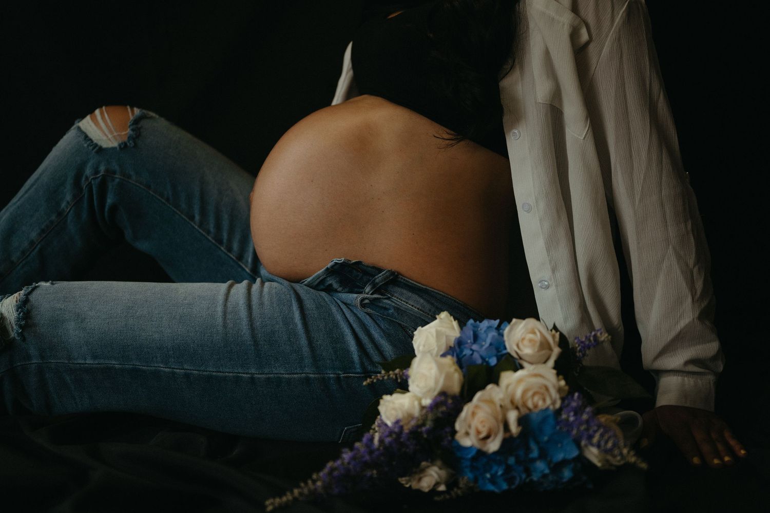 A dark moody maternity photo with white and blue roses against a black background.