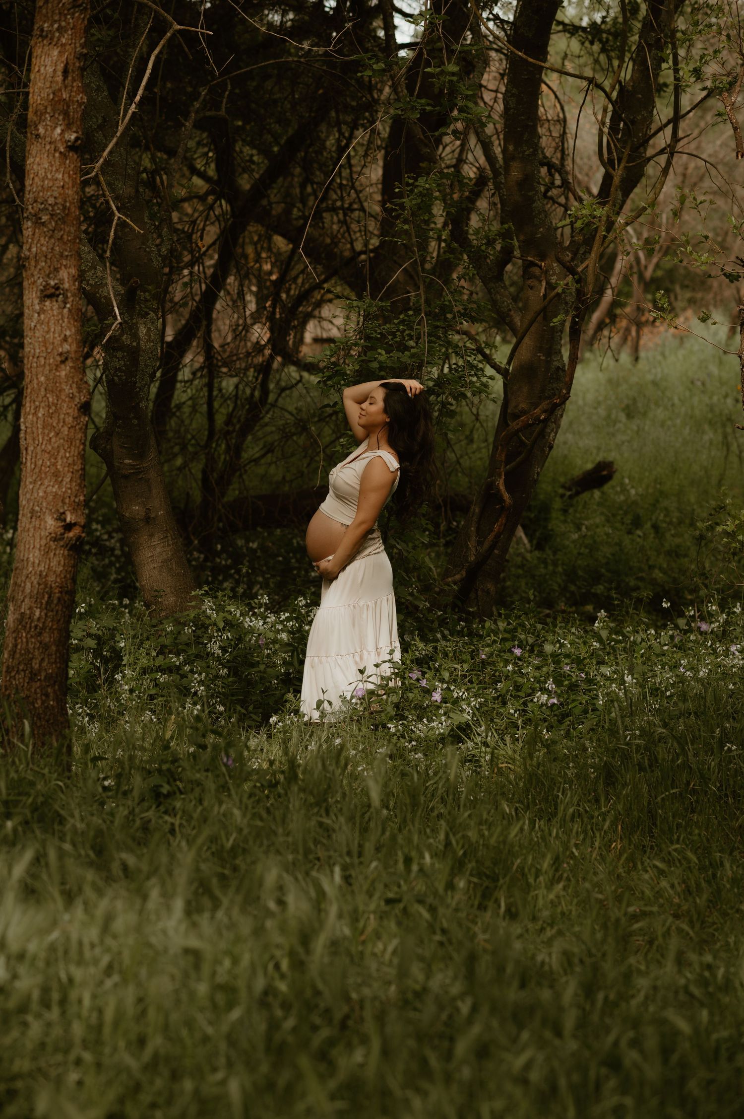 Ethereal maternity portrait in a forest with white wildflowers and moody lighting.