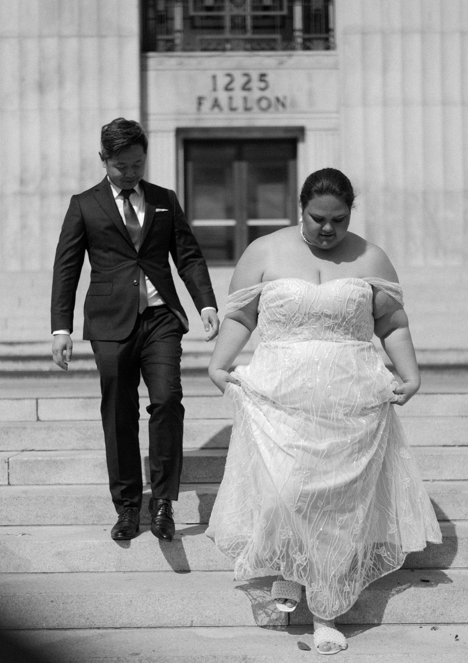 A couple in wedding attire descends stone steps outside a building marked 1225 Fallon.